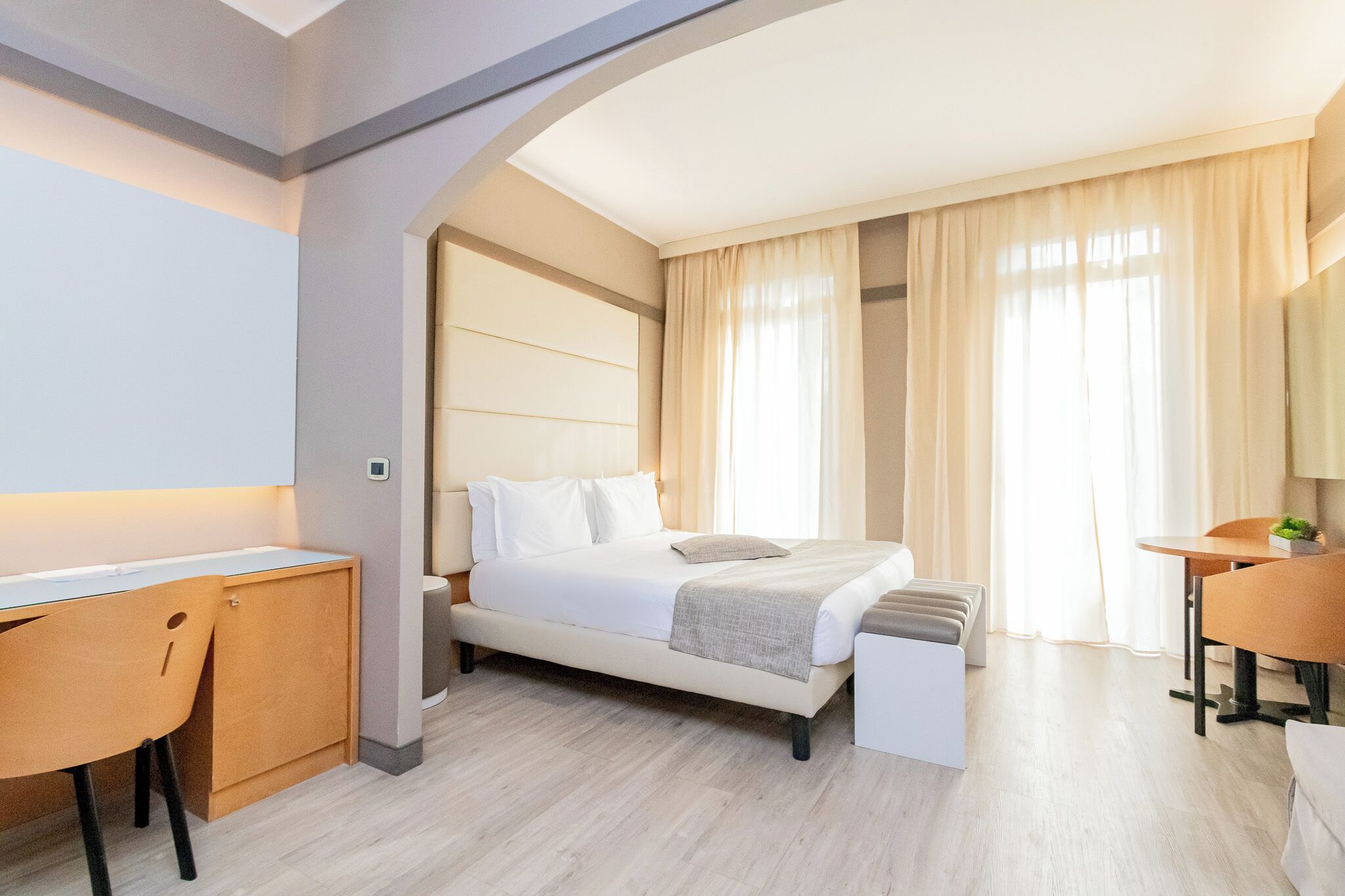 A modern hotel room with a bed, desk, and chair, a small table, and a large window with curtains.