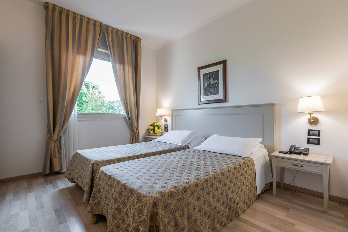 A hotel room with two beds, a desk, a lamp, a framed picture on the wall, and a window with curtains.