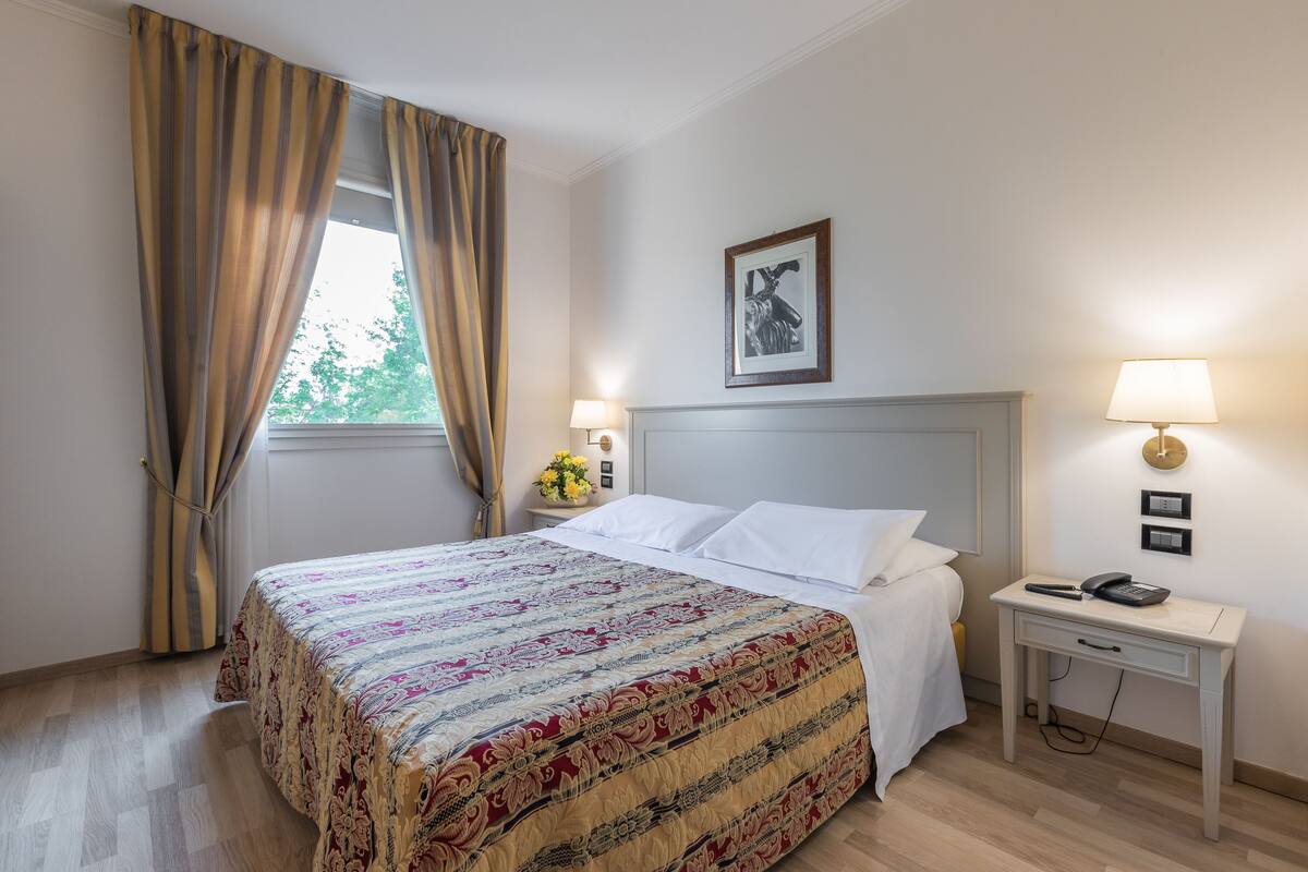 A hotel room with a bed, a nightstand with a lamp, a desk with a phone, and a window with curtains.