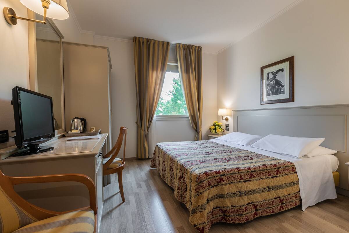 A hotel room with a bed, a desk with a computer, a chair, a bedside table with a lamp, and a window with curtains.