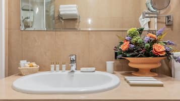 A bathroom with a white sink, a mirror, a towel rack, a flower arrangement, and toiletries on the counter.