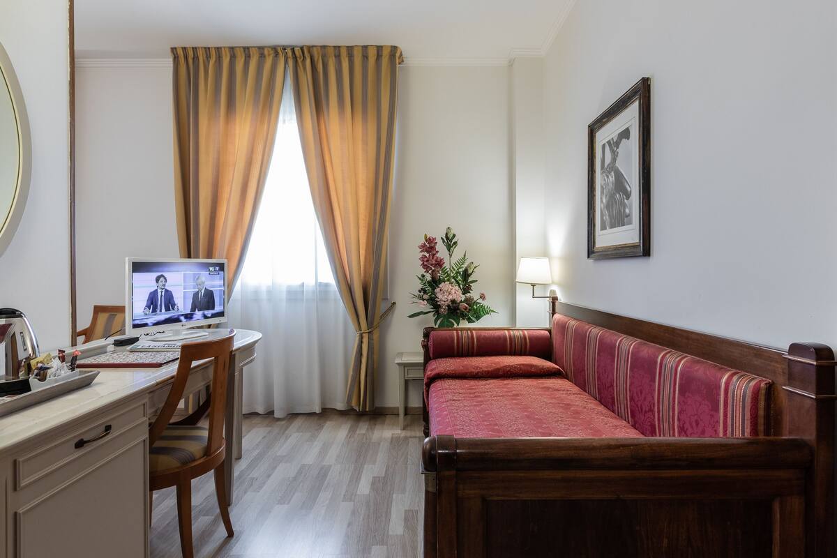A hotel room with a wooden desk, a computer, a red sofa, a framed picture on the wall, and a window with curtains.