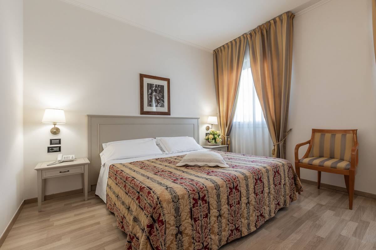 A neatly arranged hotel room with a large bed, a bedside table with a lamp, a chair, and a framed picture on the wall.