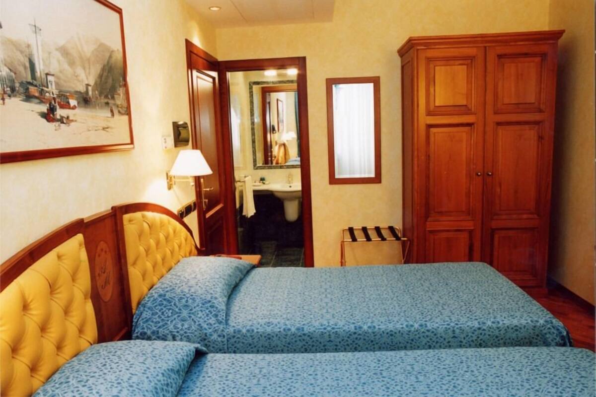A hotel room with two beds, a wooden wardrobe, a bathroom visible through an open door, and a painting on the wall.