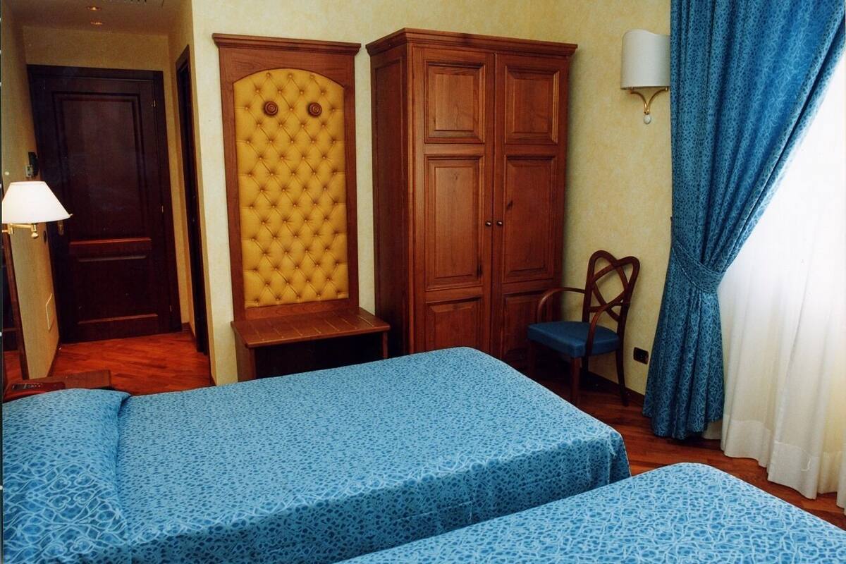 A hotel room with two beds, a wooden wardrobe, a chair, and a lamp.
