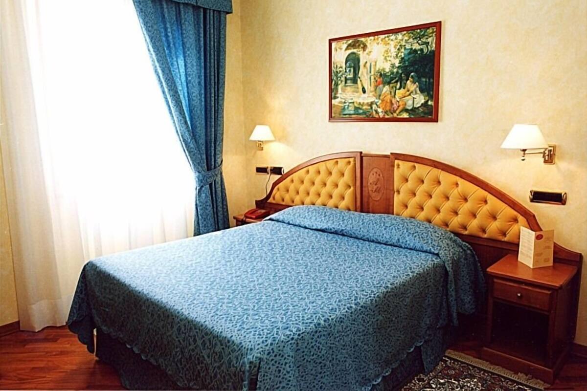 A neatly made bed with a blue bedspread, two bedside lamps, and a painting on the wall.