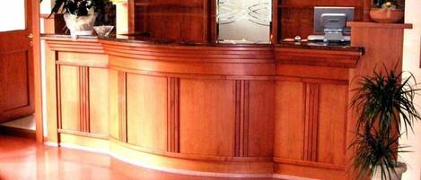 A reception area with a wooden reception desk, a large mirror, and a telephone. There are potted plants on either side of the desk.