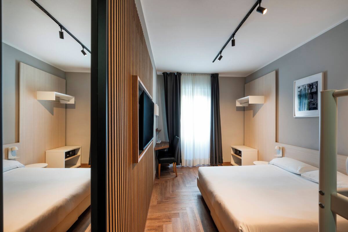 A hotel room with two beds, a TV, a desk, and a window with curtains.