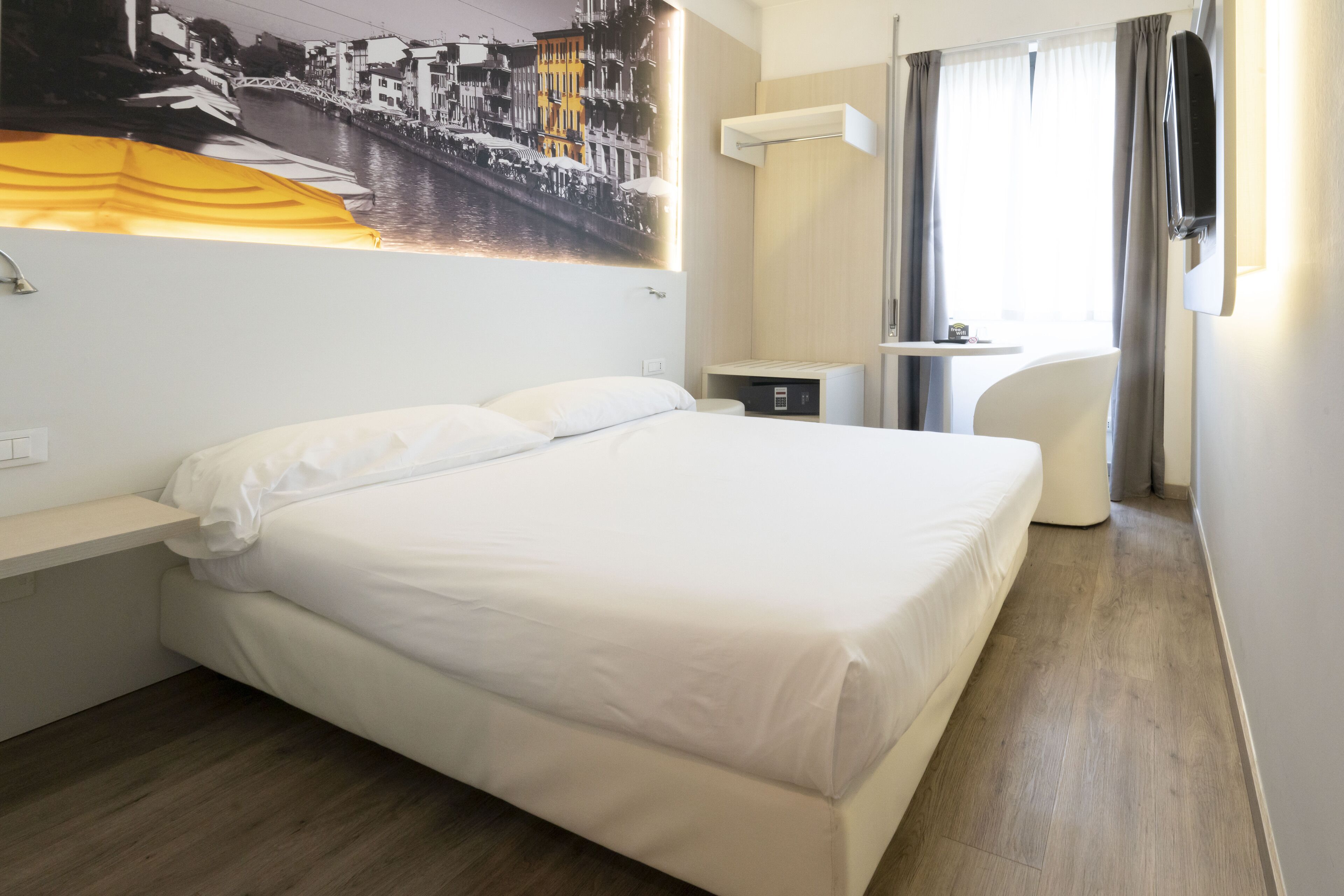 Et hotelværelse med en stor seng, et lille natbord, et vægmonteret TV og et vindue med gardiner.