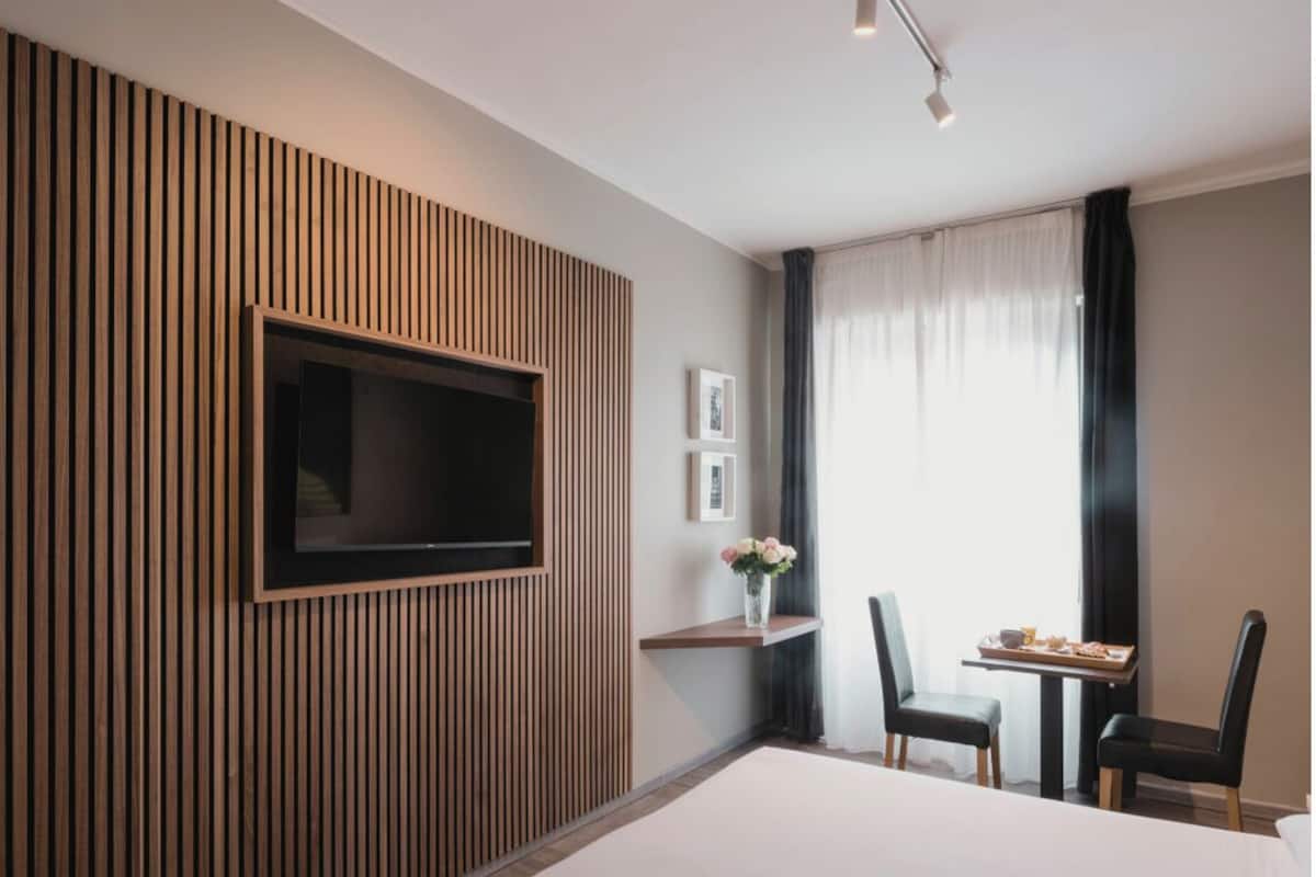 A hotel room with a bed, a TV mounted on a wooden panel wall, a small table with chairs, and a window with curtains.