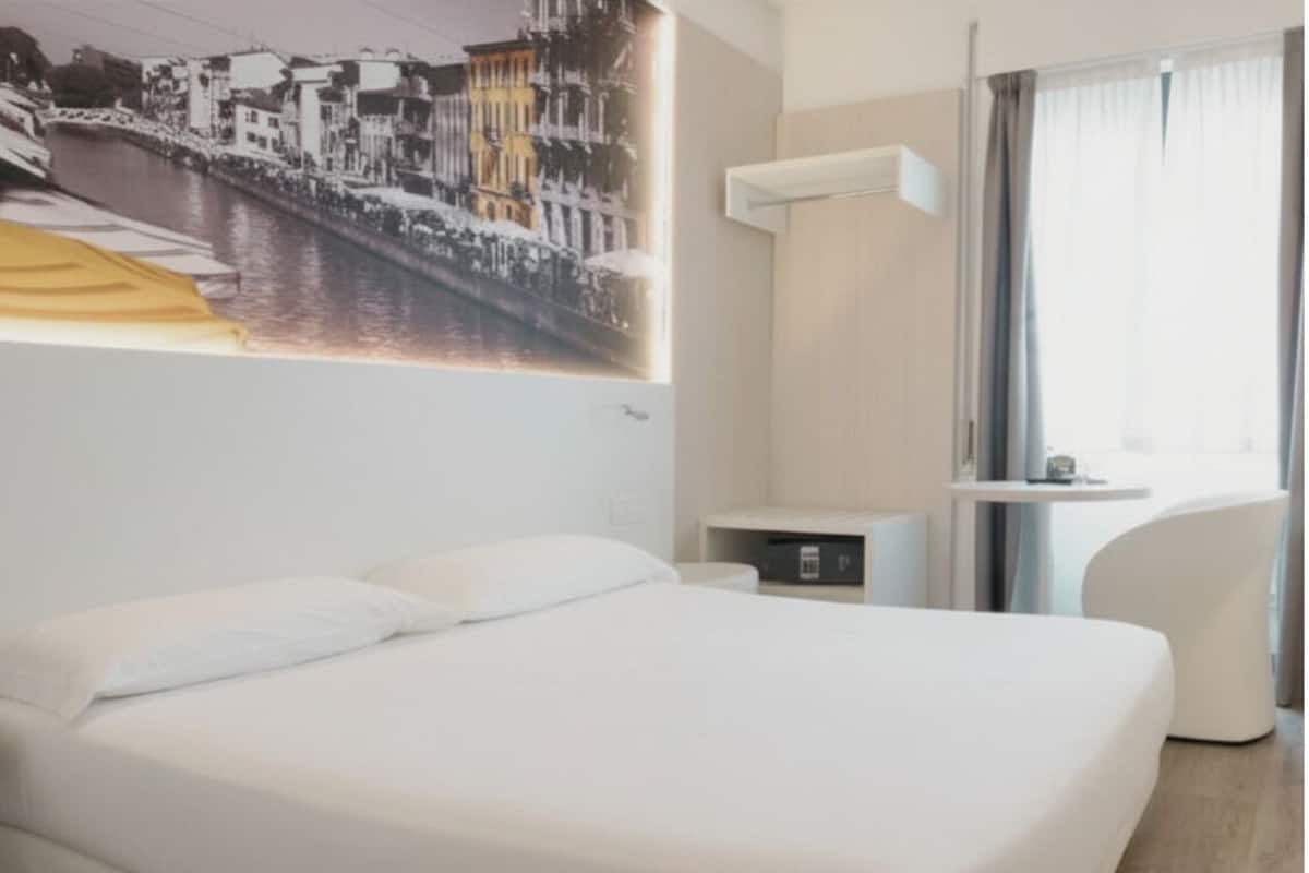 A hotel room with a bed, a desk, a chair, a window with curtains, and a wall-mounted air conditioner.