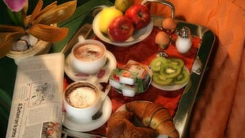 Una bandeja con cruasanes, manzanas, kiwi, huevos y dos tazas de café, además de un periódico y una planta.
