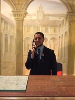 Un hombre con traje sentado a una mesa con un mapa delante, en una habitación con un gran mural de un edificio con arcos y columnas.