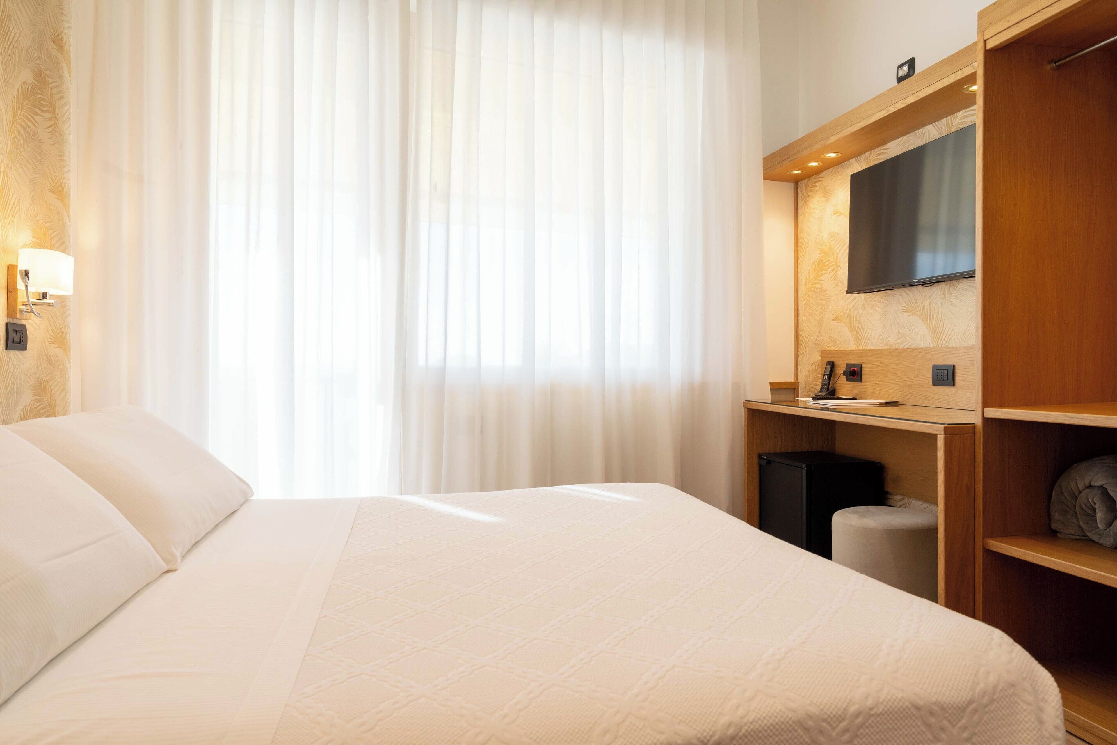 Habitación de hotel con una cama grande, un televisor de pantalla plana montado en la pared, un escritorio con una silla y un pequeño cubo de basura.