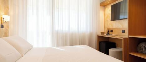 Habitación de hotel con una cama grande, un televisor de pantalla plana montado en la pared, un escritorio con una silla y un pequeño cubo de basura.