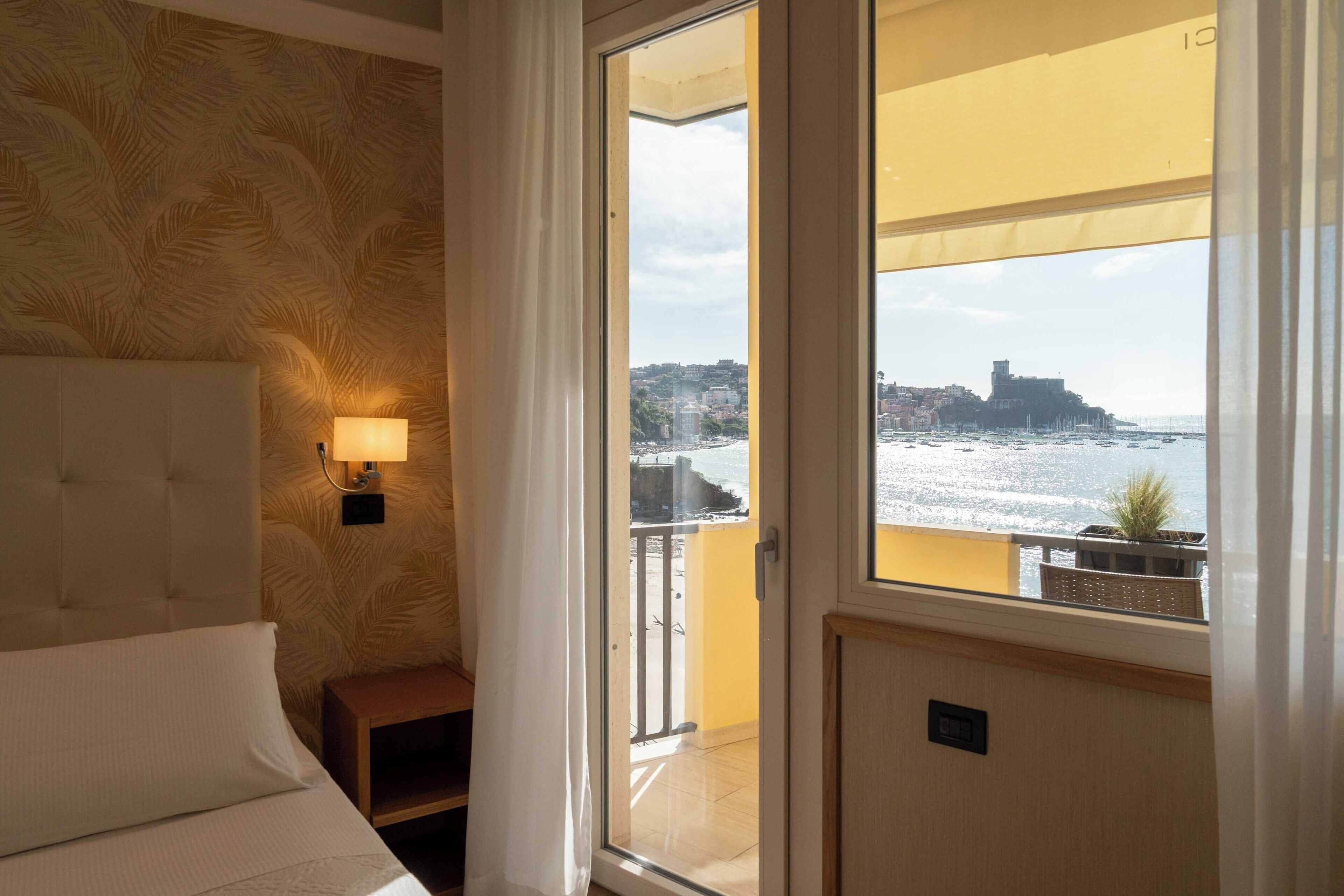 Et hotelværelse med en seng, et natbord, en lampe og en balkon med udsigt over havet og en bygning i det fjerne.