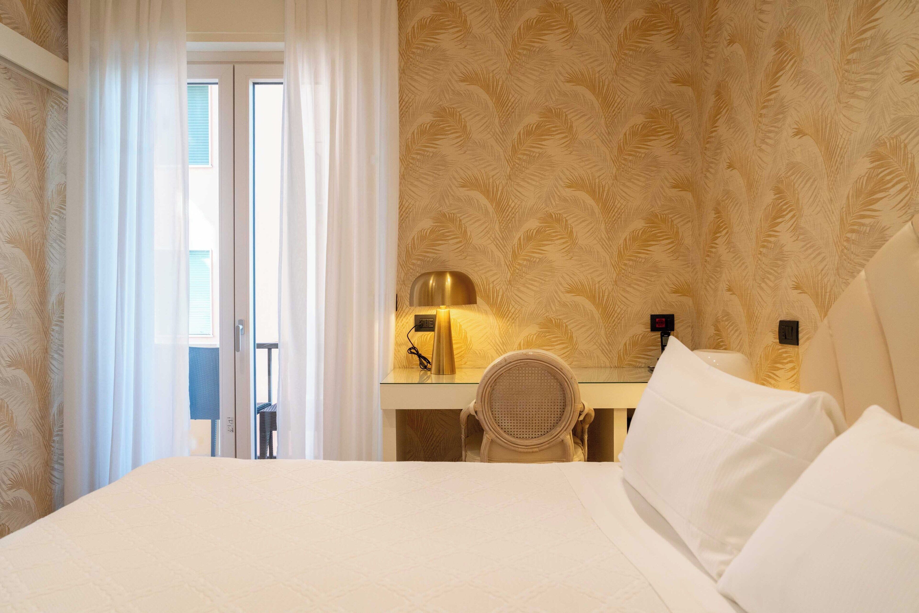 A hotel room with a bed, a desk with a lamp, a chair, and a view of the outside through a door.