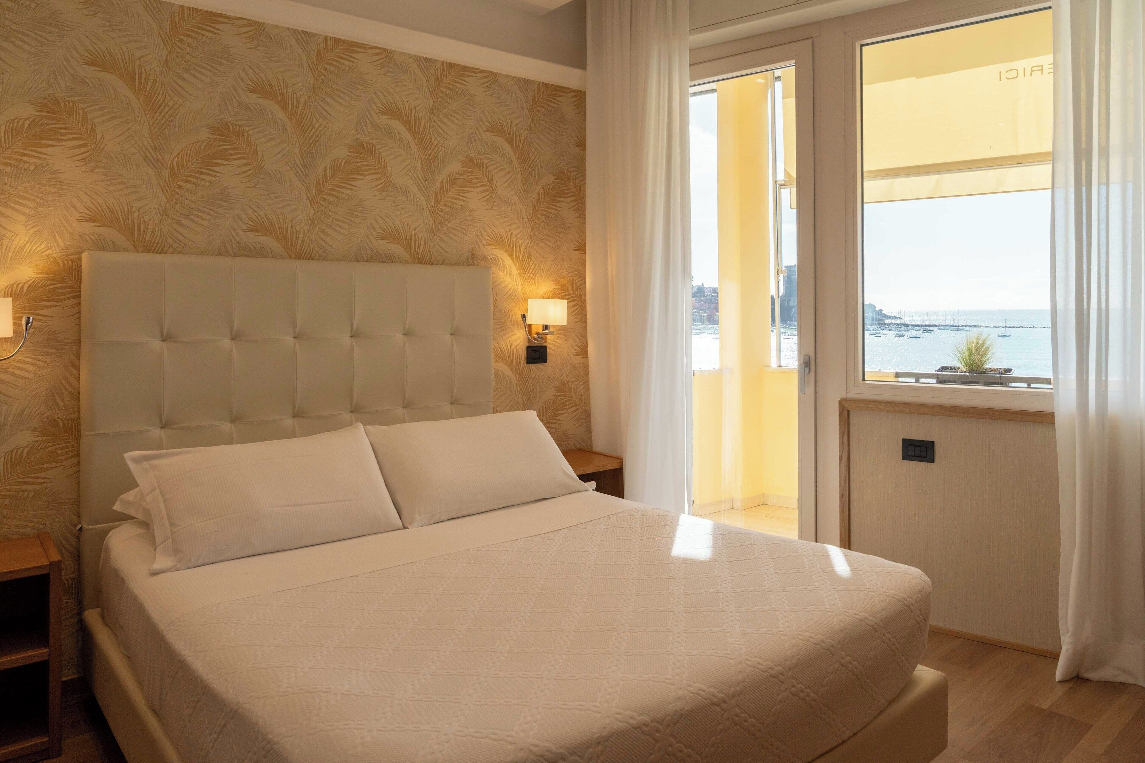 A hotel room with a large bed, a headboard with a light fixture, a nightstand, and a view of the sea through a window.
