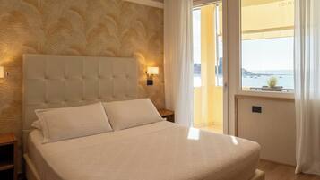 A hotel room with a large bed, a headboard with a light fixture, a nightstand, and a view of the sea through a window.