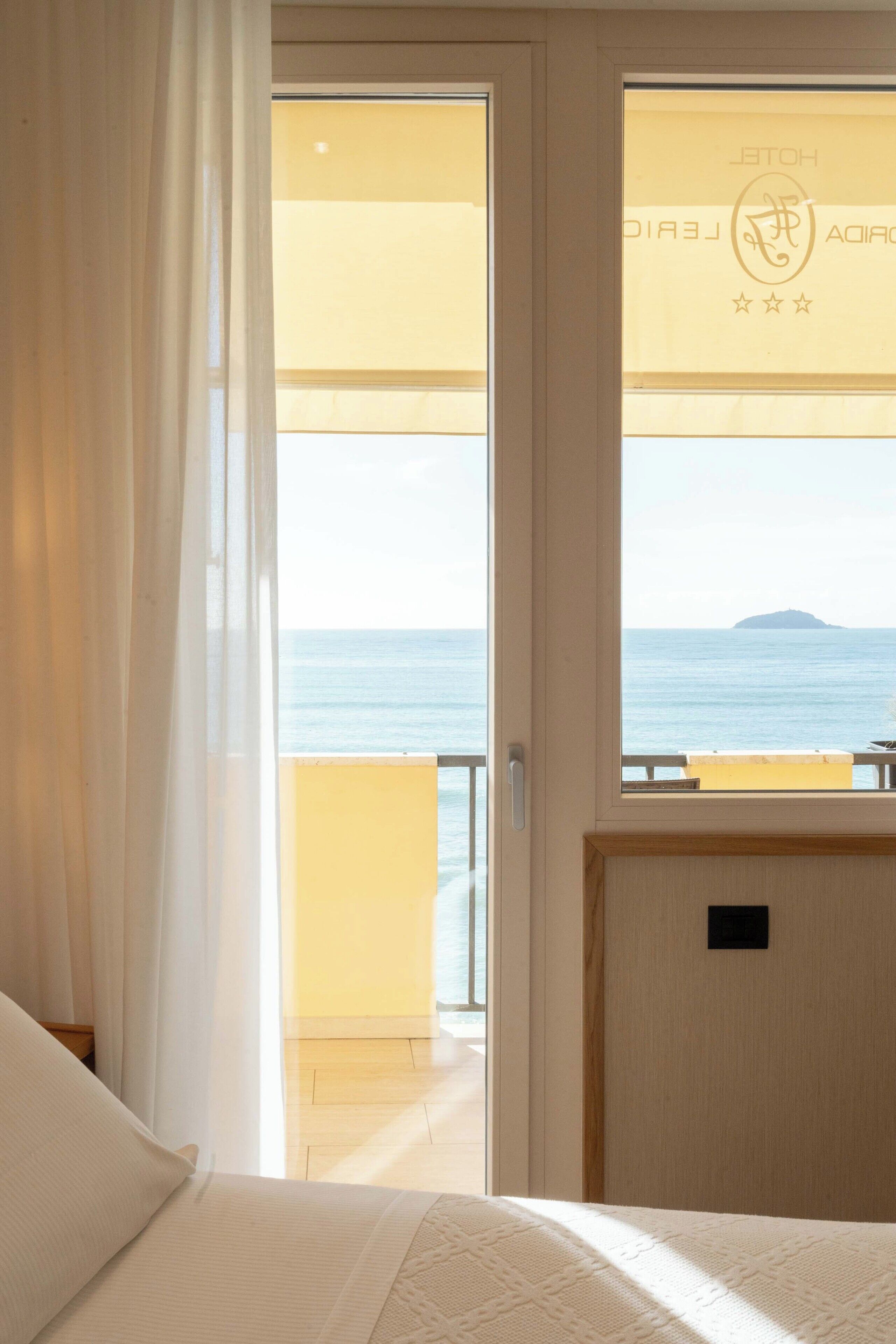 Habitación de hotel con cama, ventana con vista al mar y balcón con barandilla amarilla.