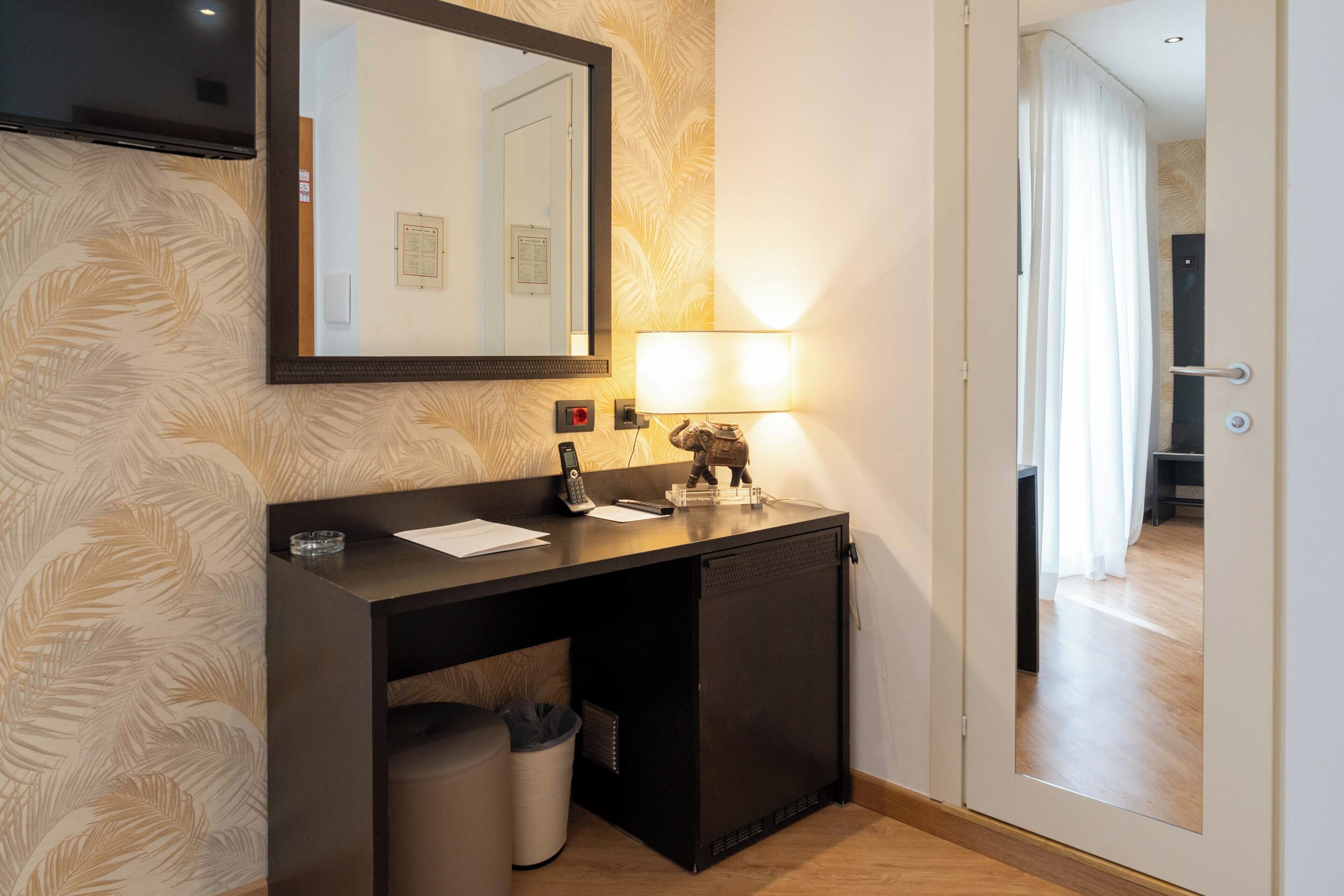 A hotel room with a dark wooden desk, a mirror, a wall-mounted telephone, a small trash bin, and a lit wall lamp.