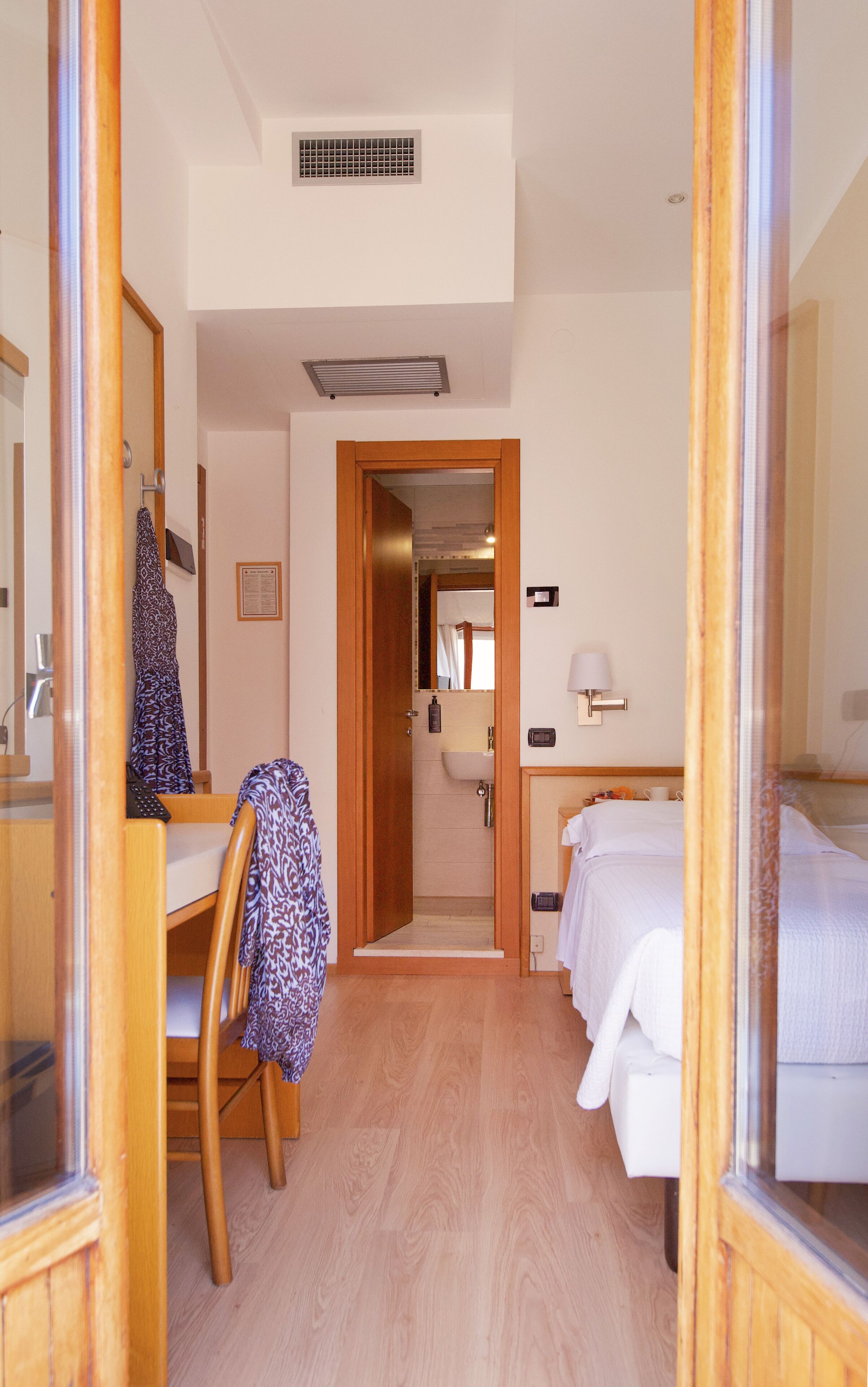 Hotelkamer met houten vloer, bureau met stoel, bed, nachtkastje, spiegel, raam en een zichtbare badkamer door een open deur.