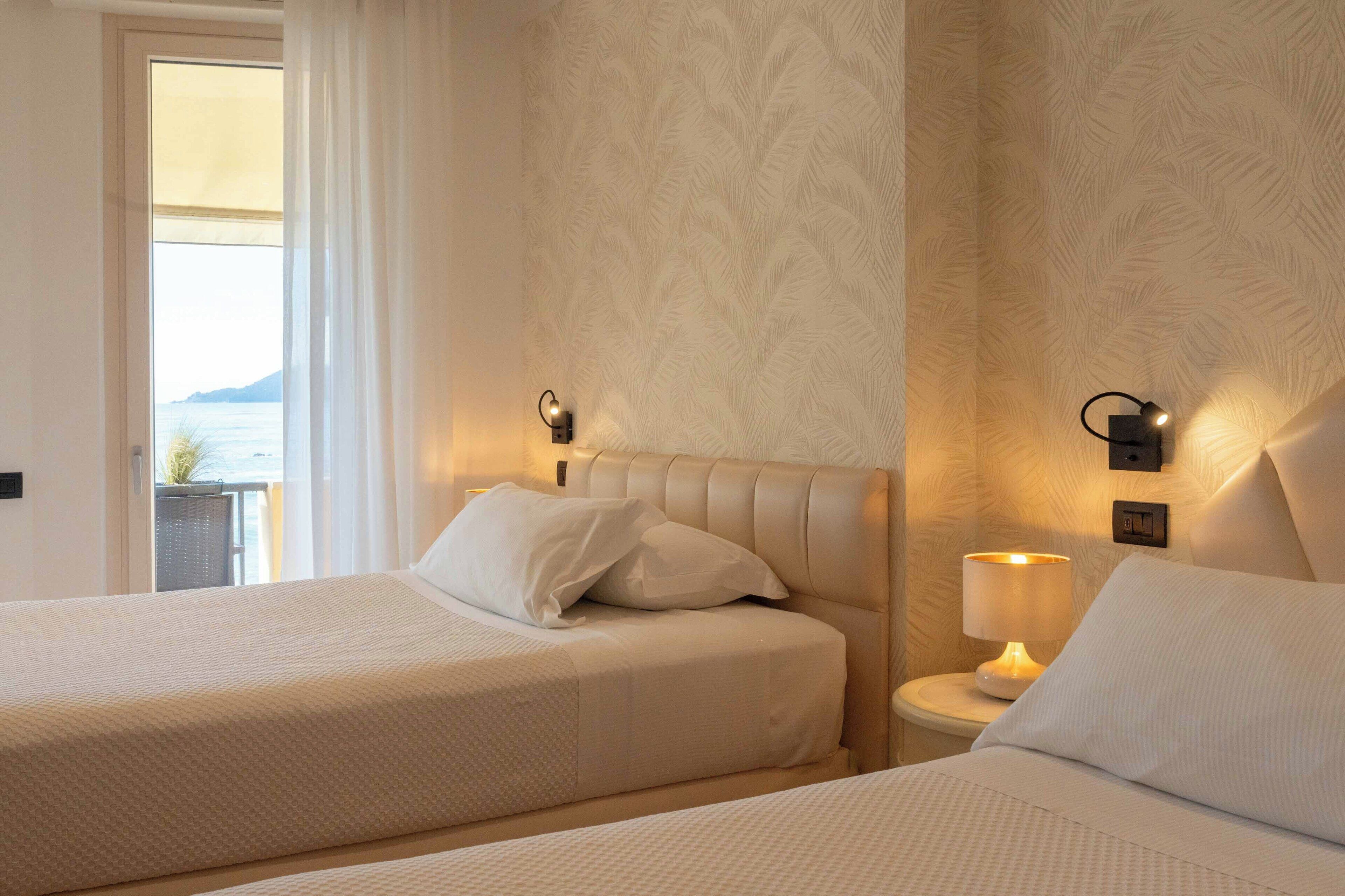 Habitación de hotel con dos camas, una mesita de noche con lámpara y vistas al mar a través de una ventana.