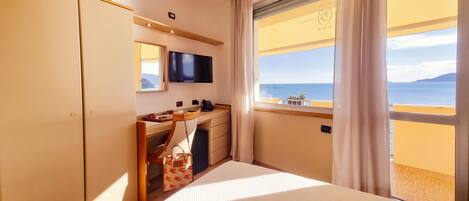 A hotel room with a bed, a desk with a chair, a television, and a window with a view of the sea.