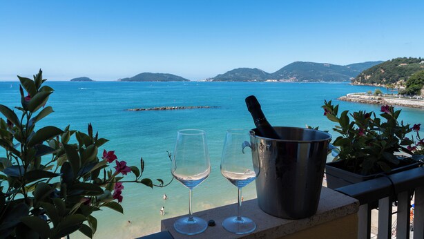 Ein Balkon mit zwei Weingläsern und einer Champagnerflasche, mit Blick auf das klare, blaue Meer und die entfernten Hügel.
