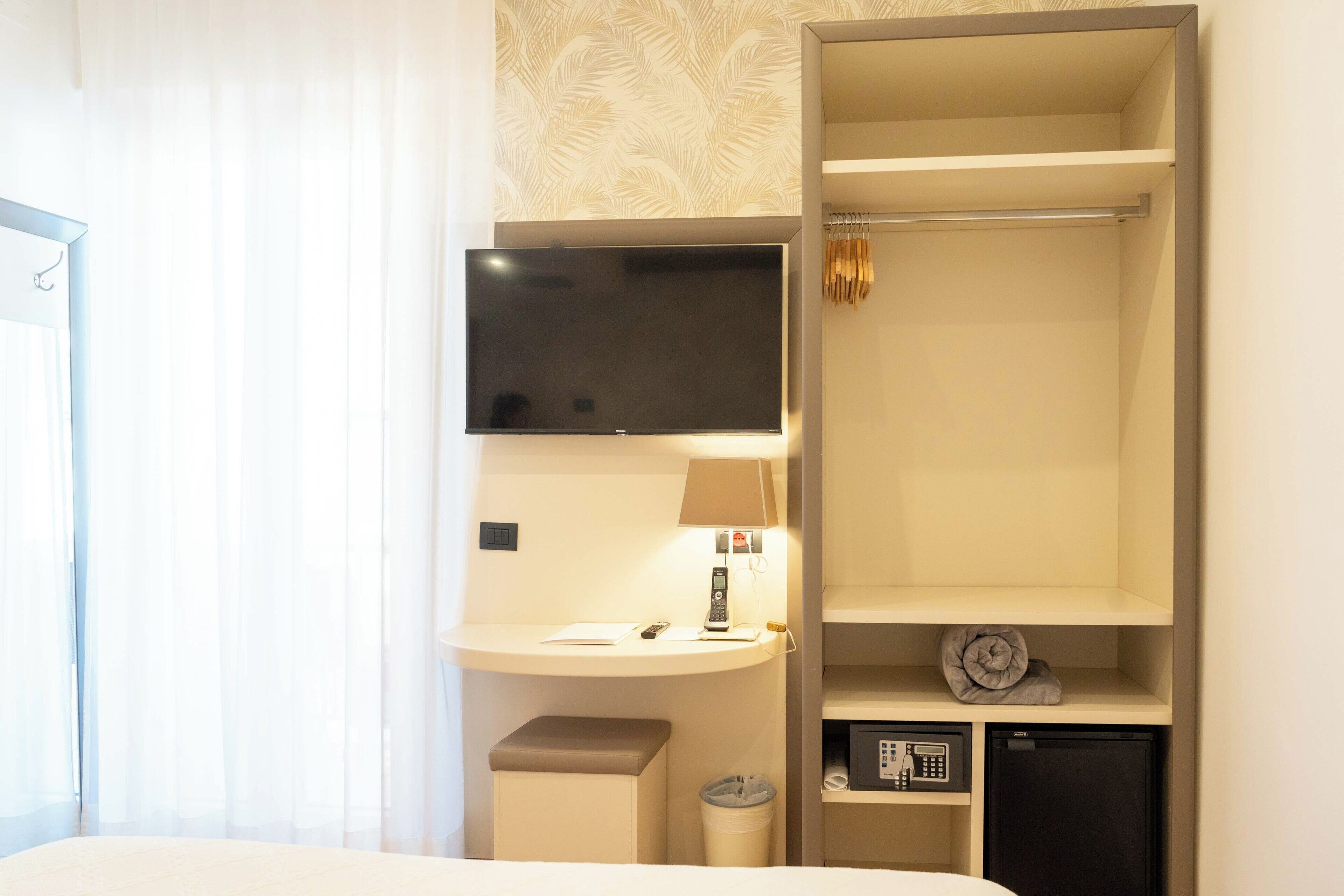 A hotel room with a bed, a desk with a lamp, a TV, a telephone, a small safe, and a minibar.
