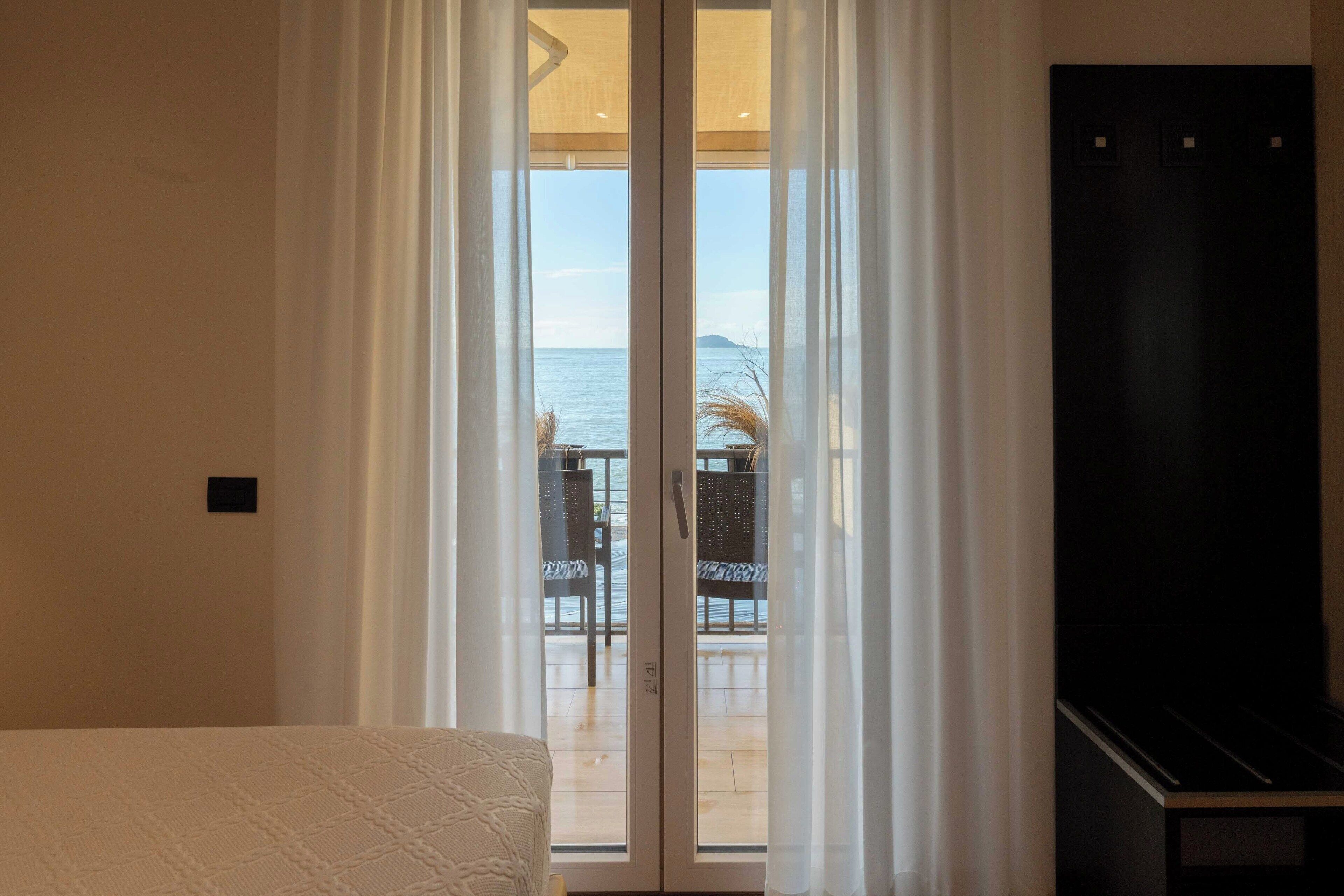 A room with a bed, a TV, and a sliding glass door leading to a balcony with a view of the sea.