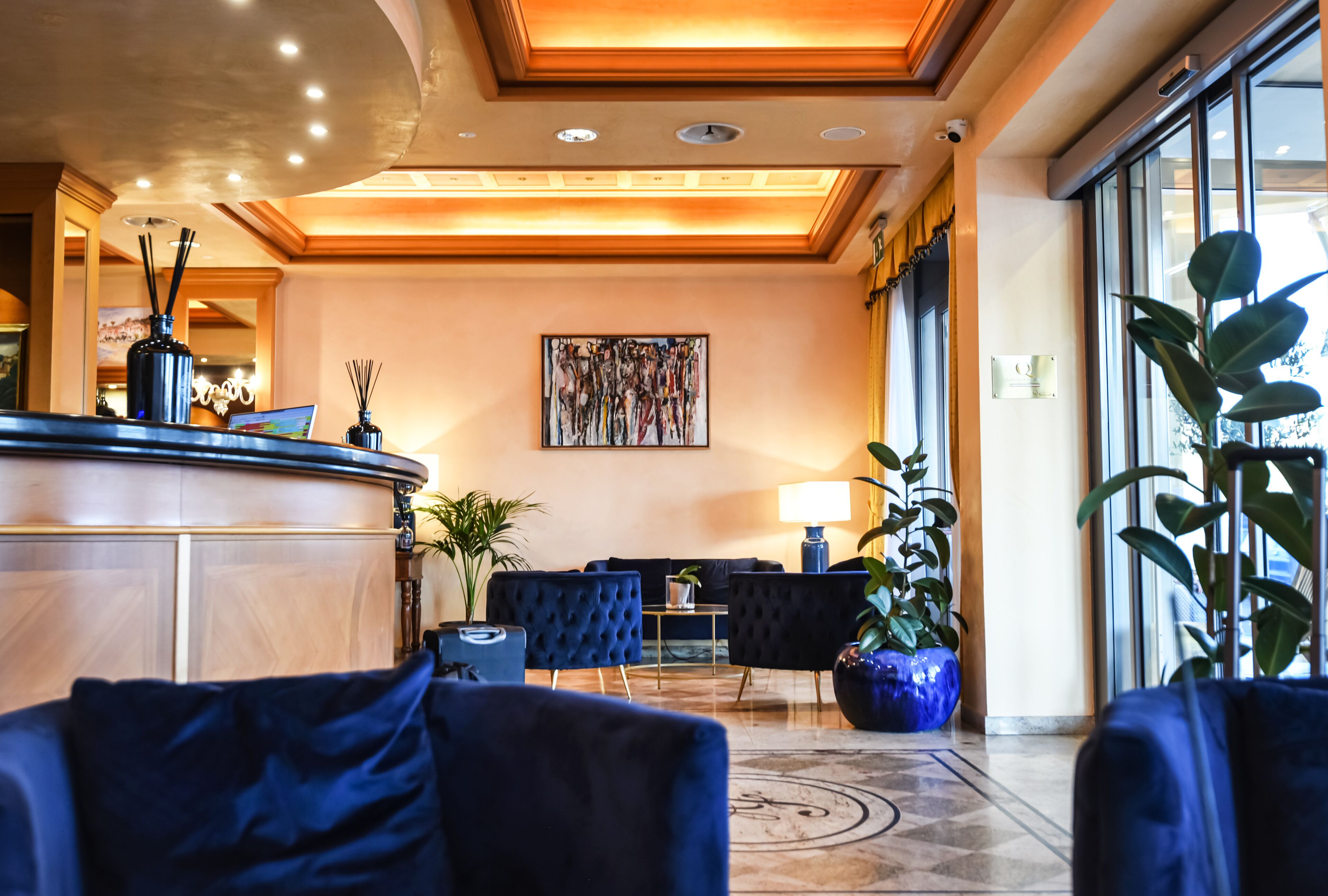 Un vestíbulo de hotel con una recepción, sofás azul oscuro, un cuadro abstracto grande y plantas en macetas.