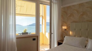 Ein Hotelzimmer mit Bett, Nachttisch, Lampe und Meerblick durch ein offenes Fenster.