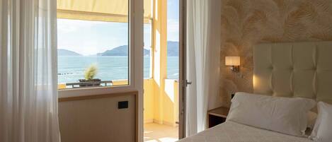 Habitación de hotel con cama, mesita de noche, lámpara y vista al mar por una ventana abierta.