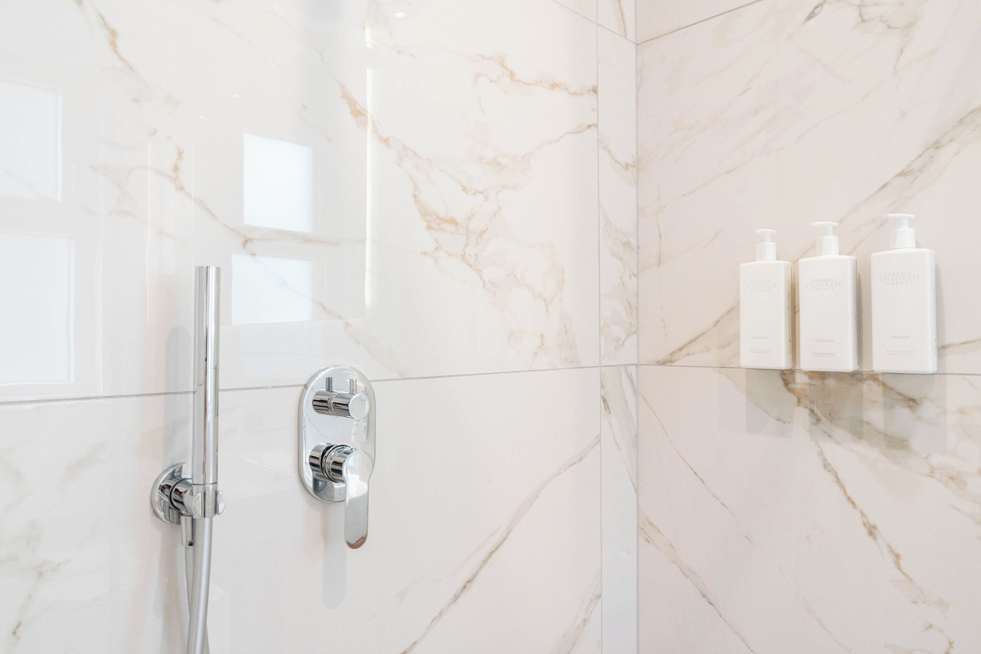 A bathroom with a marble wall, a shower, and three white bottles on the wall.