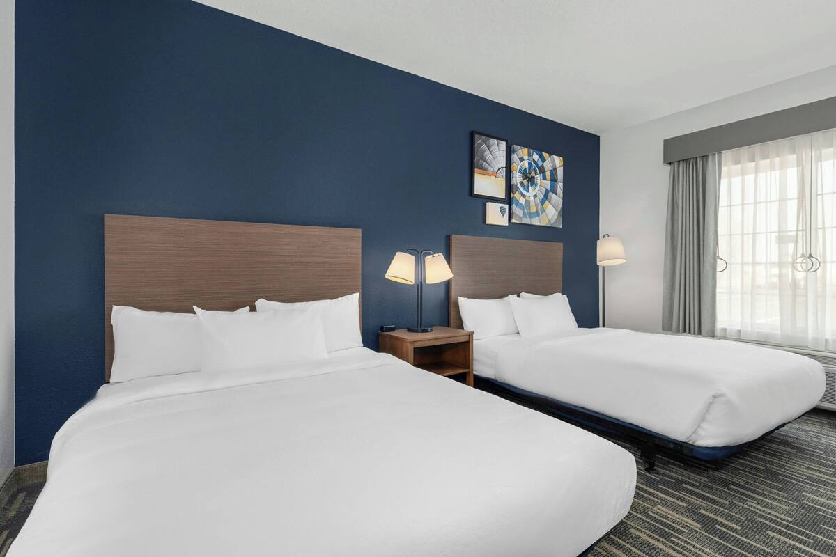 A hotel room with two beds, a nightstand with lamps, artwork on the wall, and a window with curtains.