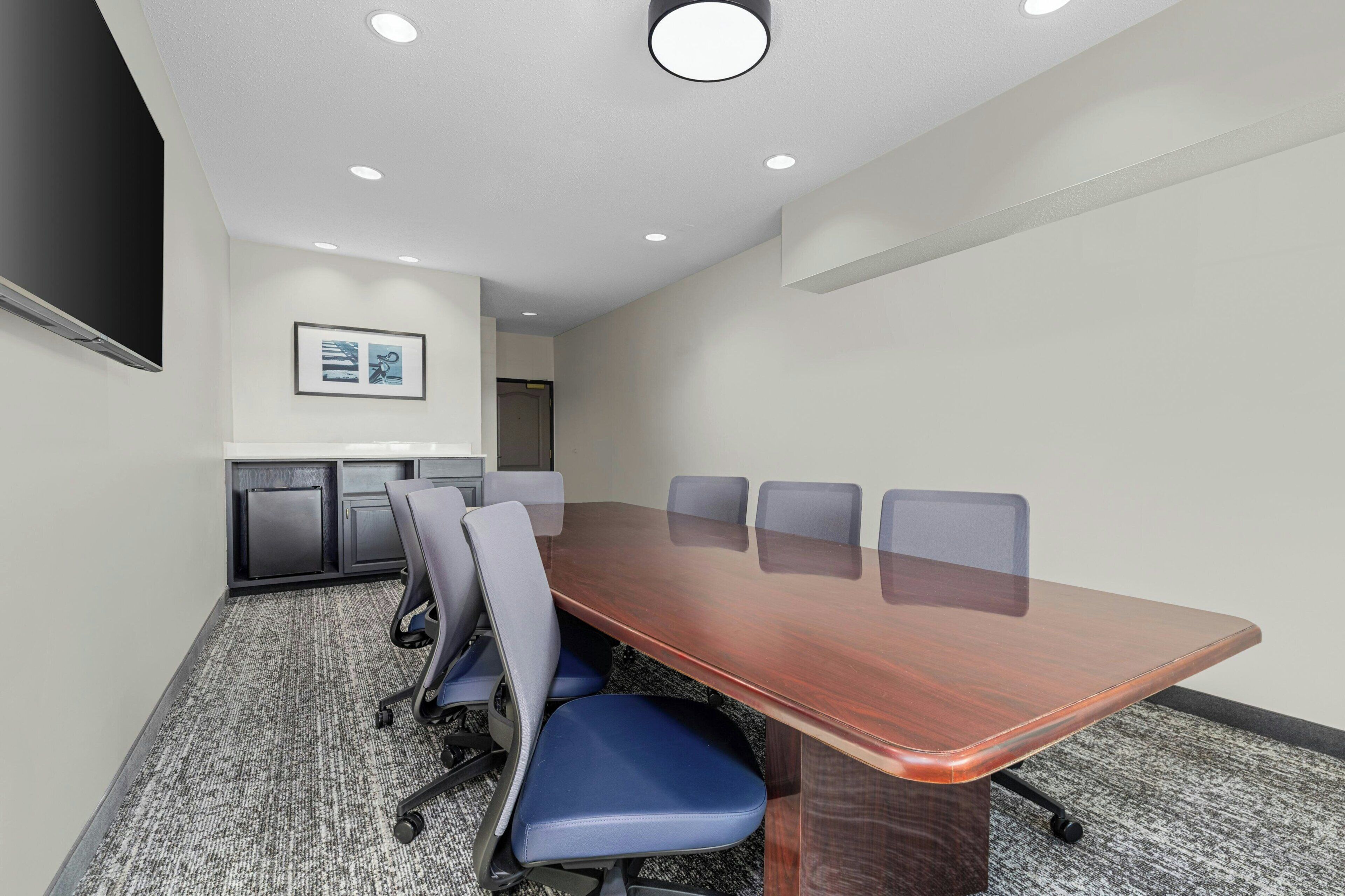 A conference room with a long table, six chairs, a projector screen, and a framed picture on the wall.