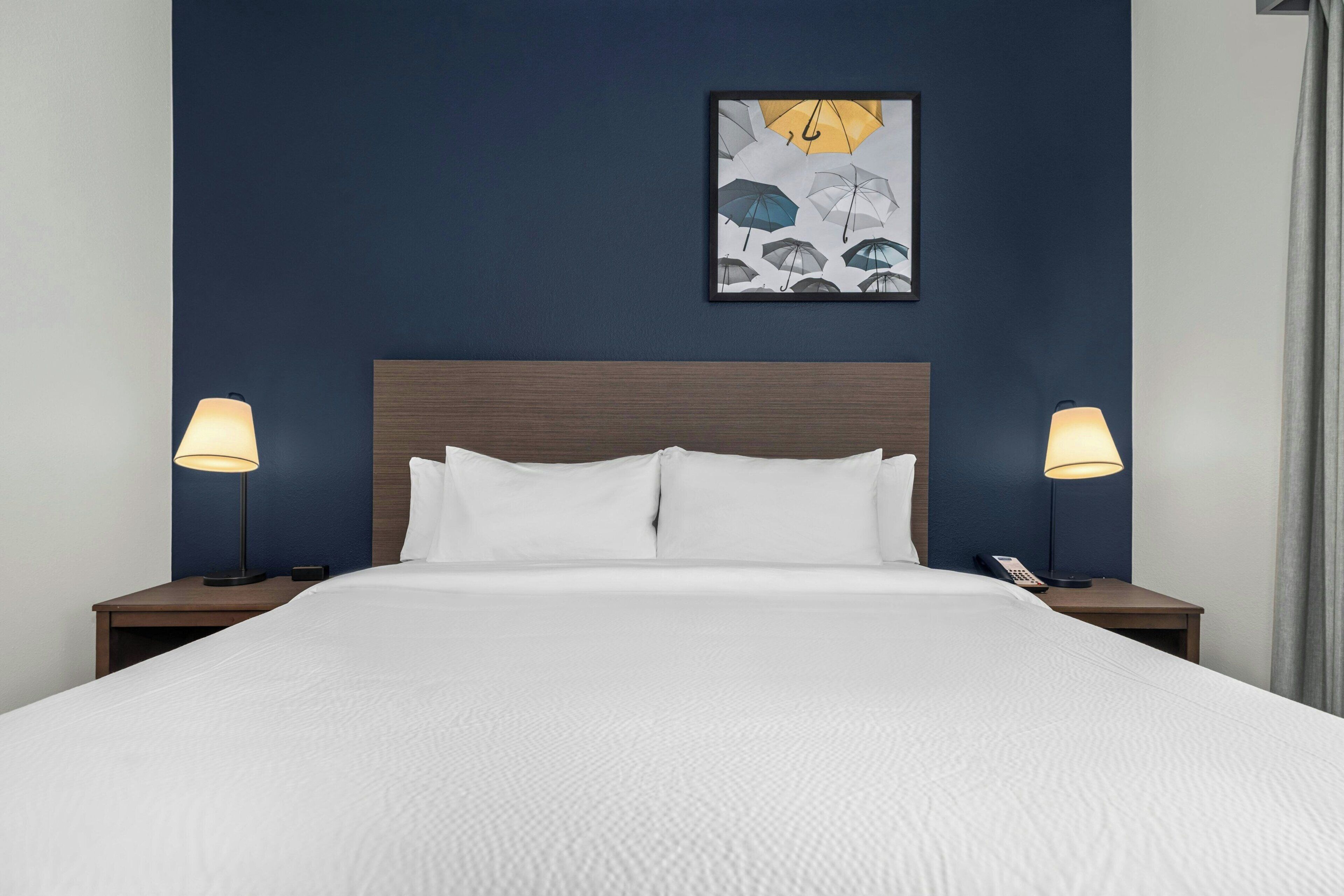 A neatly made bed with white linens, two bedside lamps, a framed artwork above the bed, and a wooden headboard.