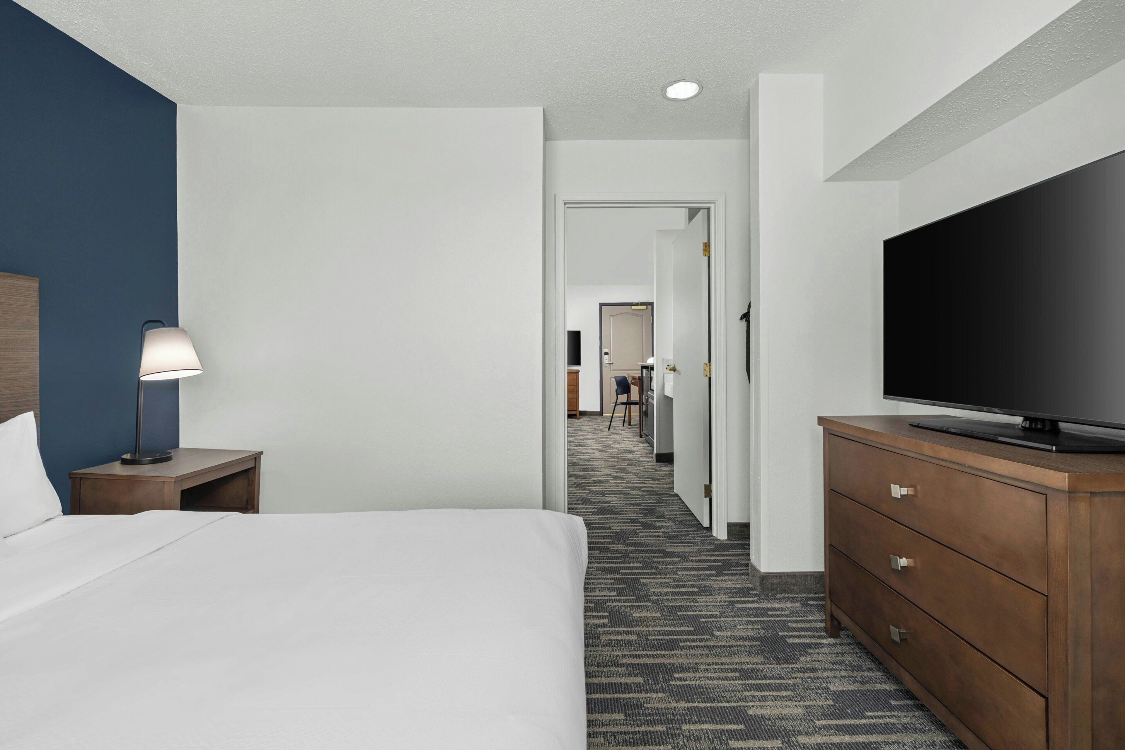 A hotel room with a bed, a TV, a nightstand with a lamp, and a dresser.