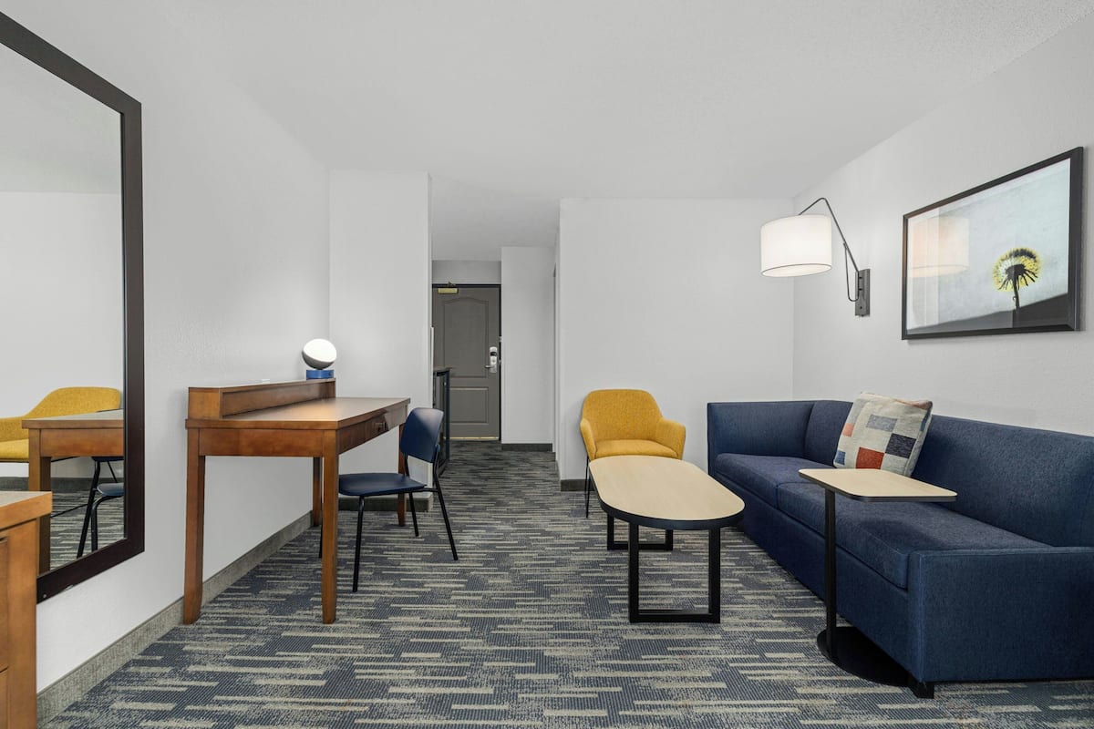 A hotel room with a desk, two chairs, a sofa, a small table, a lamp, and a framed picture on the wall.