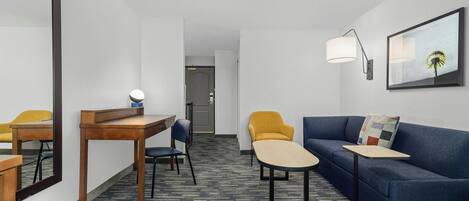 Ett hotellrum med ett skrivbord, två stolar, en soffa, ett litet bord, en lampa och en inramad tavla på väggen.
