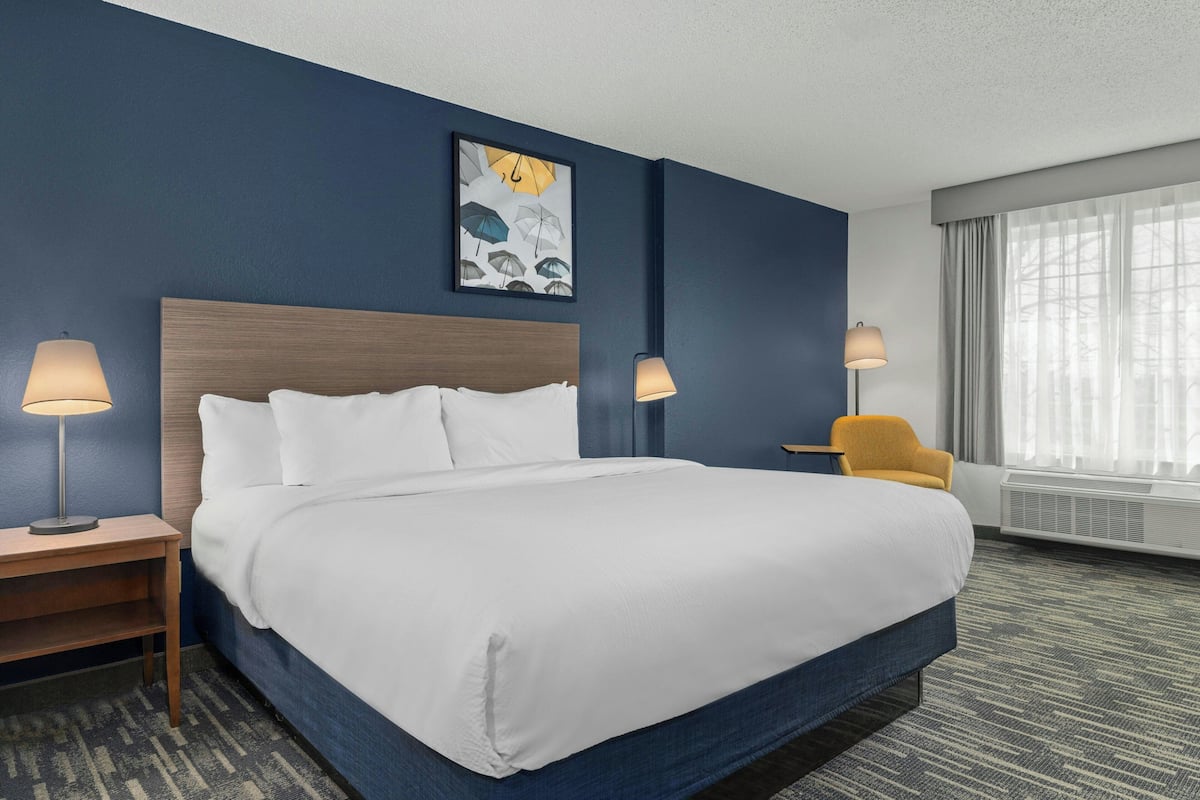 A hotel room with a large bed, two bedside tables with lamps, a chair, a window with curtains, and a wall-mounted artwork.