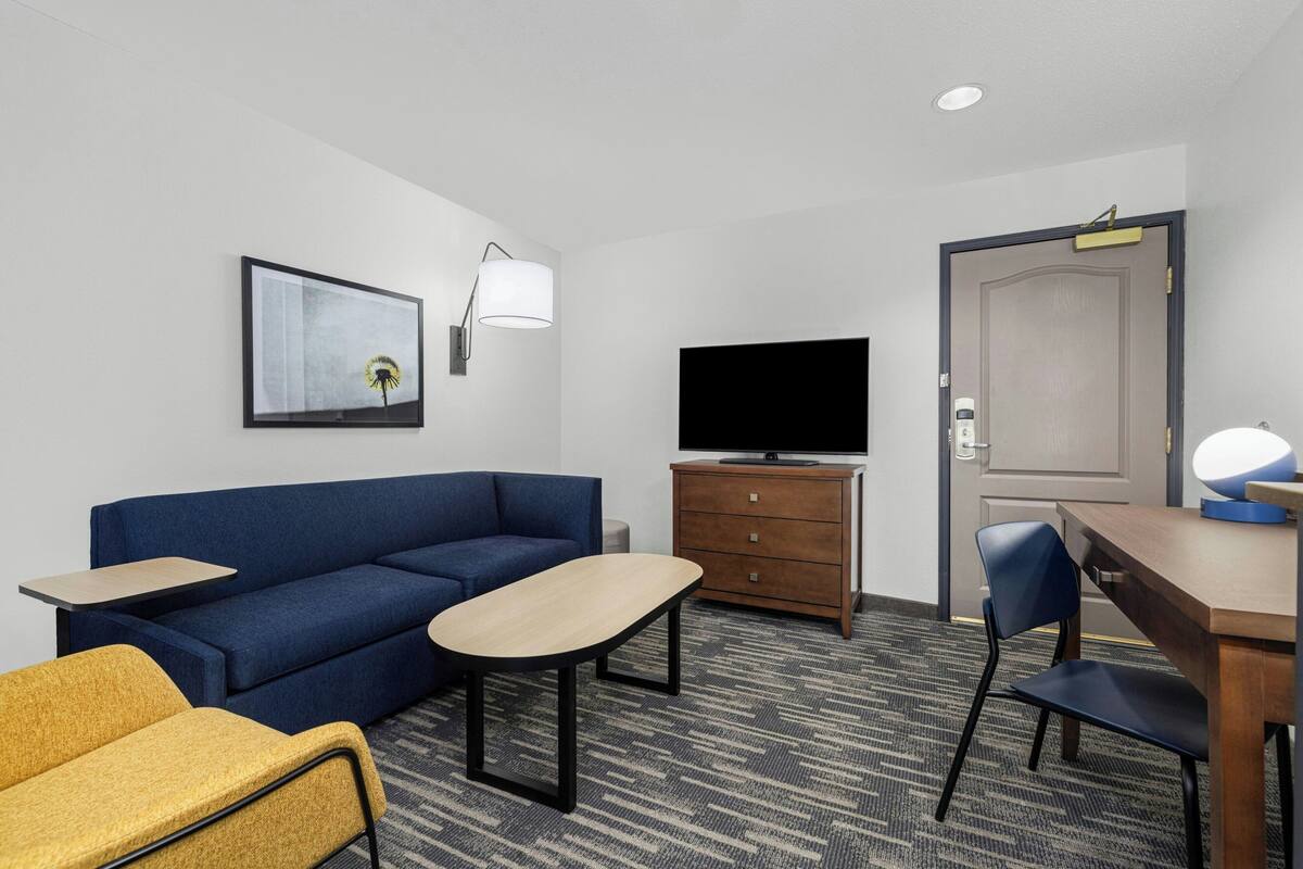 A hotel room with a blue sofa, a wooden dresser, a television, a desk with a lamp, and a chair.