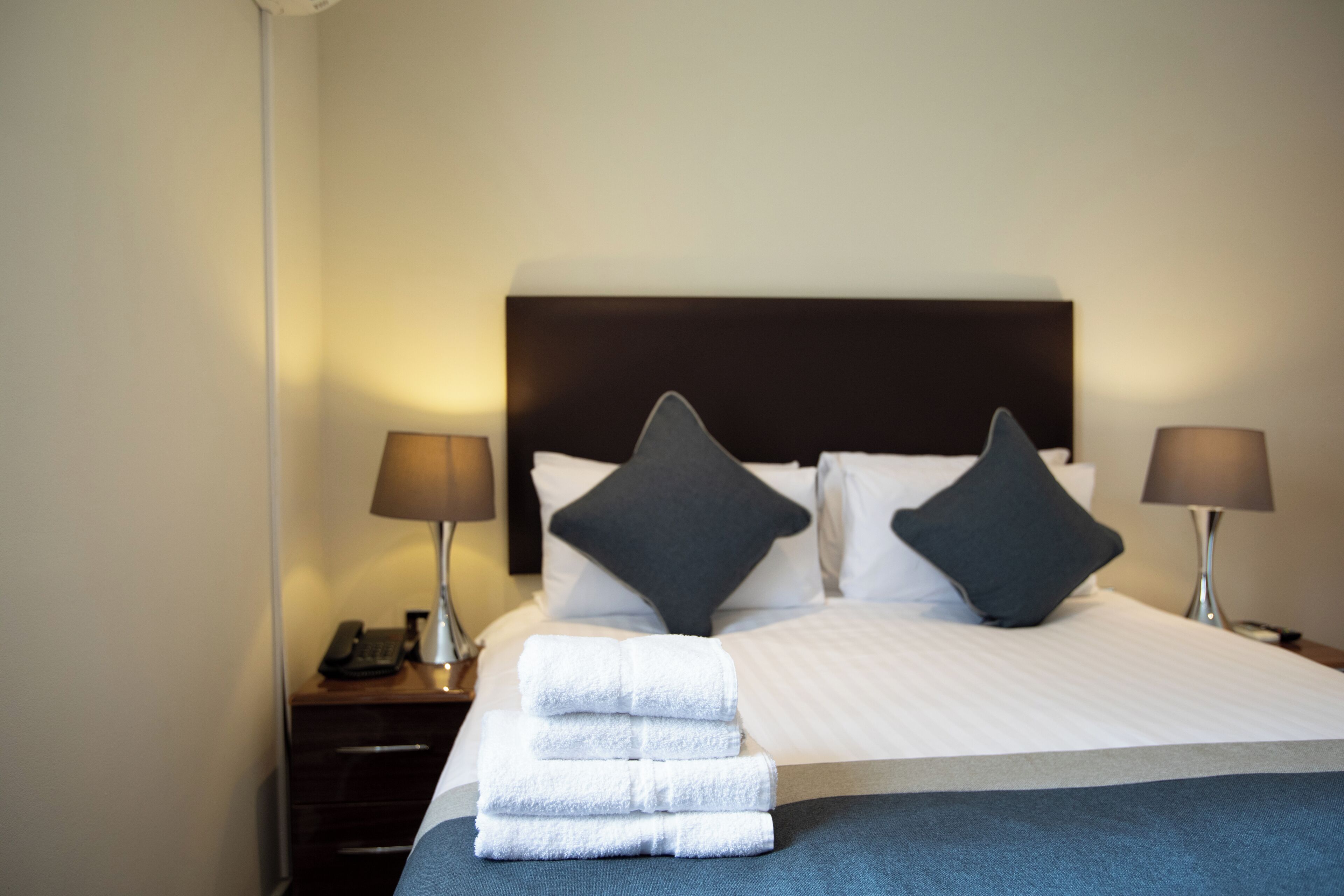 Una cama bien hecha con sábanas blancas, dos almohadas oscuras y una pila de toallas blancas sobre la cama. Hay dos mesitas de noche, una de ellas con una lámpara y un teléfono.