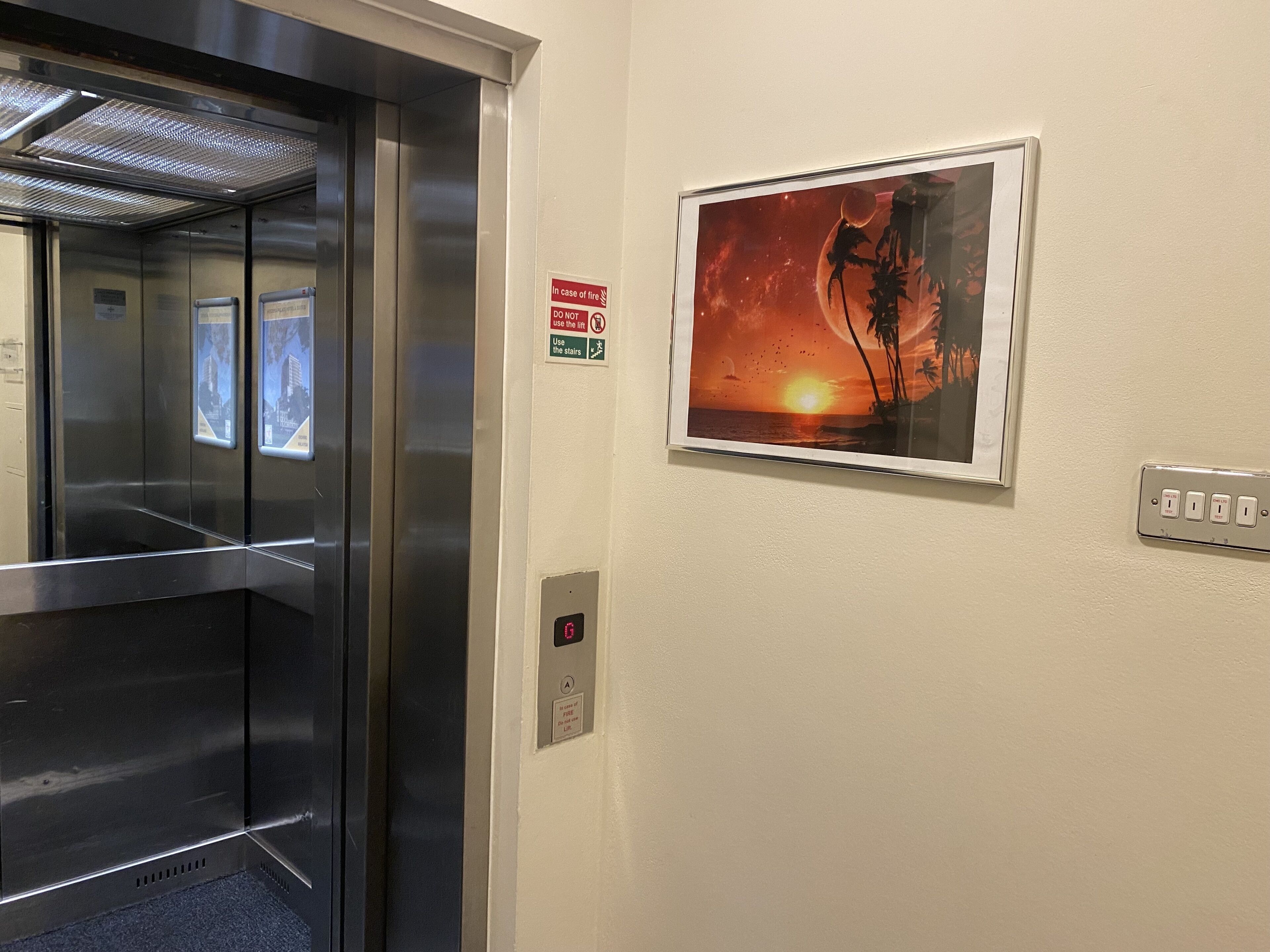 L’intérieur d’un ascenseur avec des portes en acier inoxydable, un panneau d’indication concernant la sécurité incendie, une photographie encadrée représentant un coucher de soleil et une prise électrique fixée au mur.
