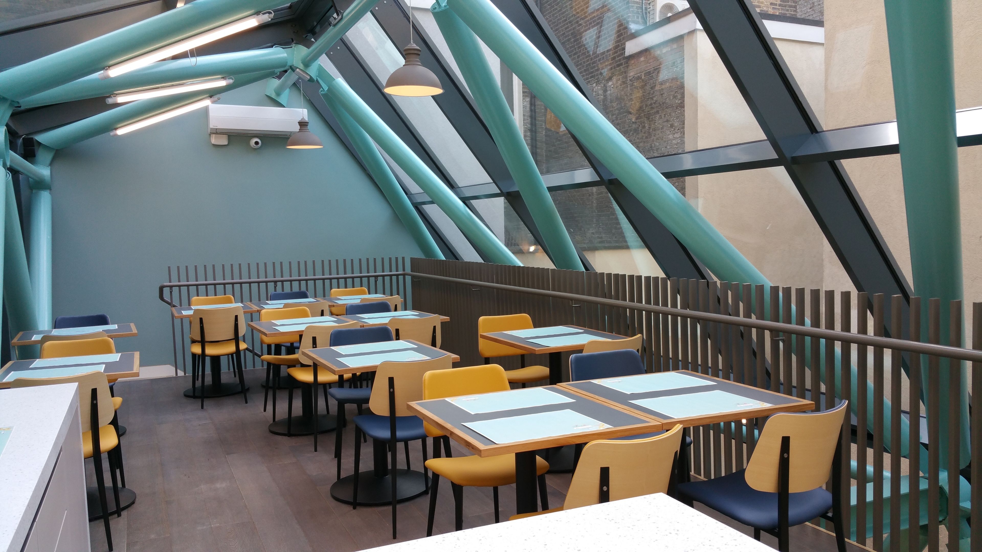 Un restaurant moderne doté d’un plafond haut et de poutres apparentes. Les tables et les chaises sont disposées dans un espace vaste et aéré.