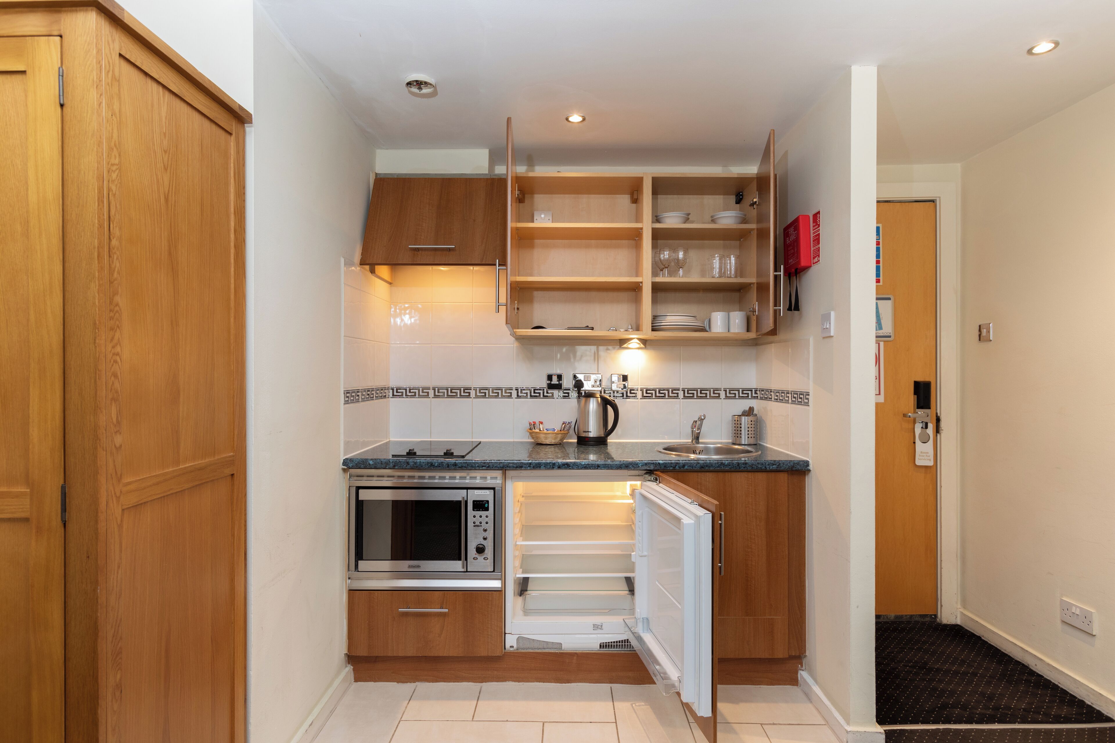 Eine kleine Küche mit Holzausstattung, einem integrierten Mikrowellenherd, einem Kühlschrank und einer Arbeitsplatte mit Wasserkocher und Tassen.