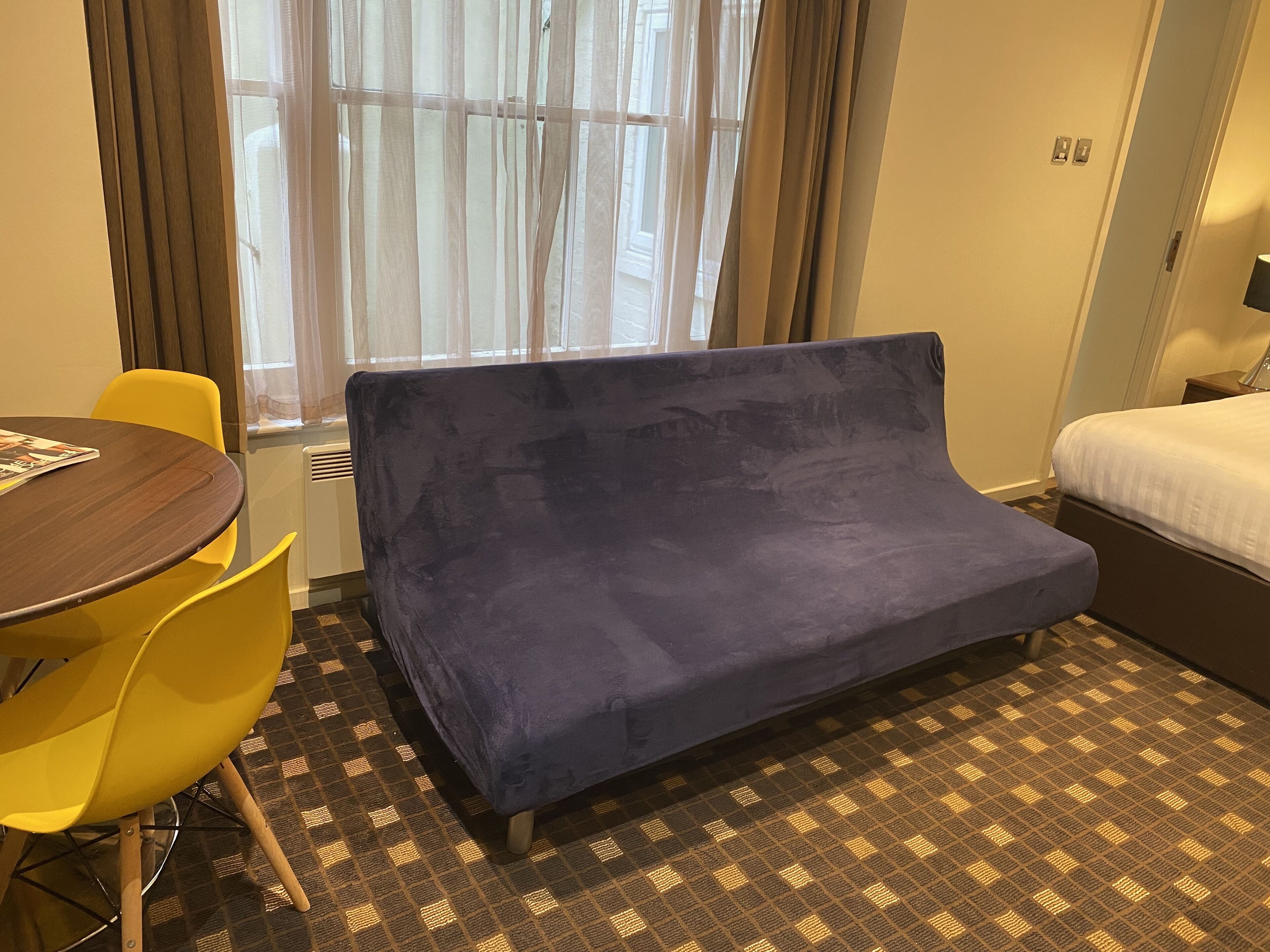 A hotel room with a blue sofa, yellow chairs, a wooden table, and a bed with a white mattress.