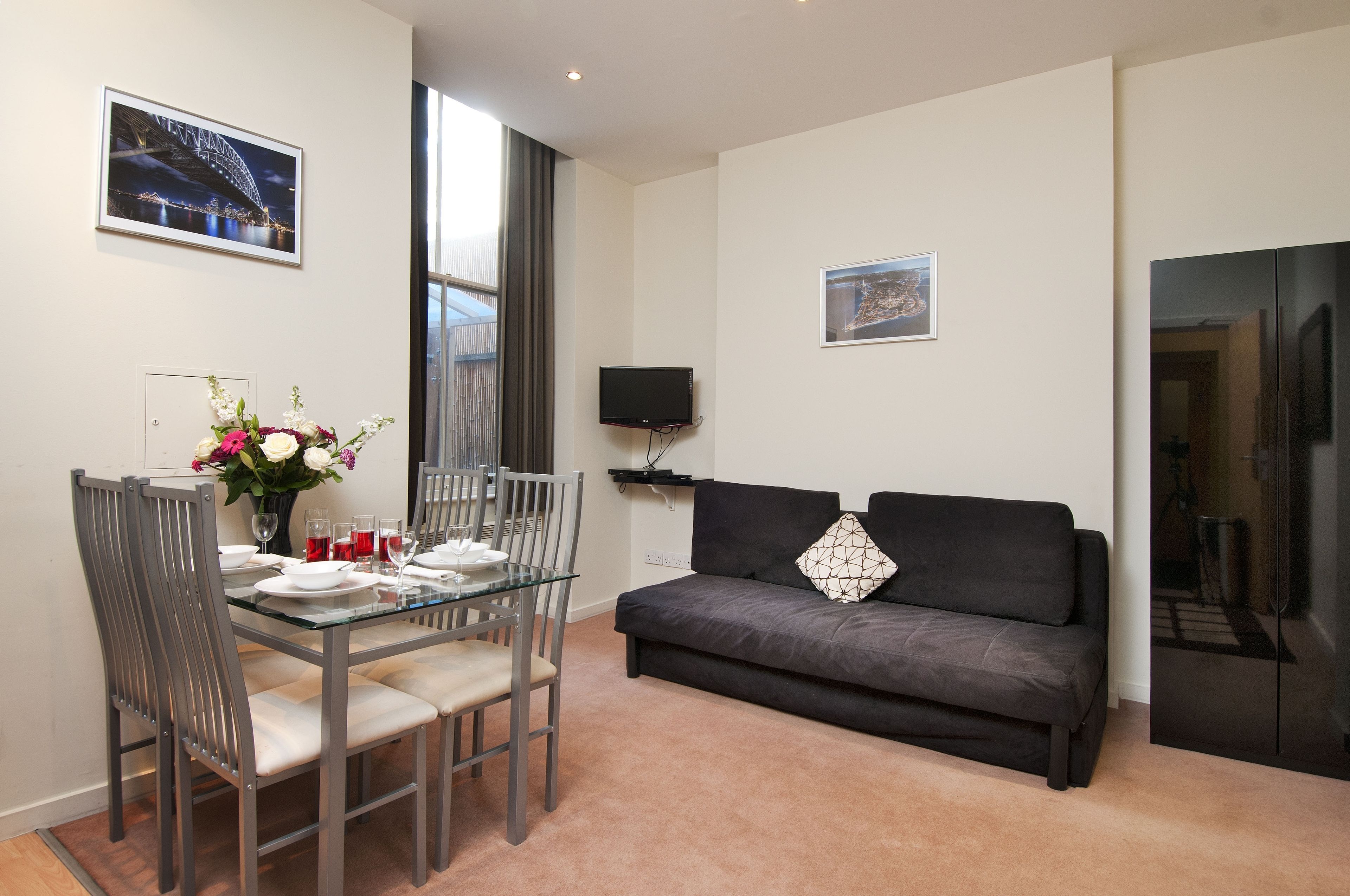 A modern living room with a glass dining table set for four, a dark grey sofa with a decorative pillow, a flat-screen TV mounted on the wall, and framed pictures on the walls.