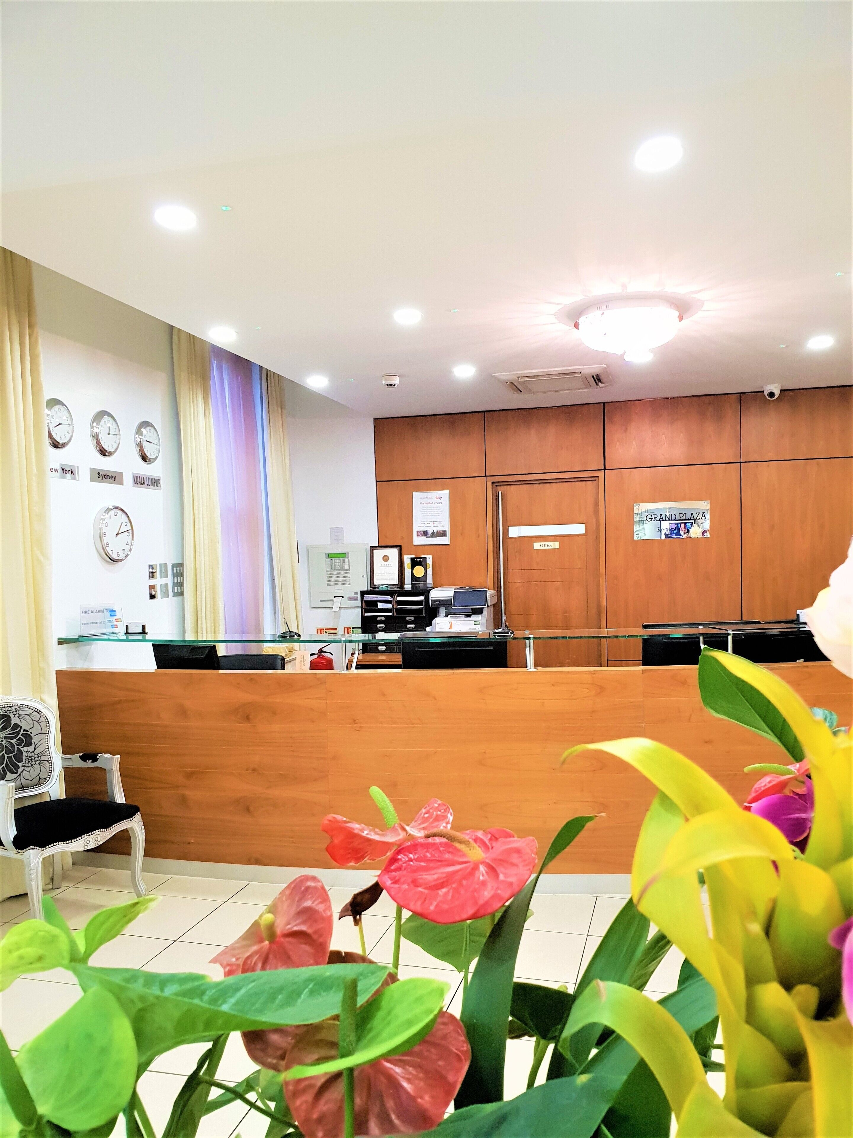 A reception area with a wooden counter, a fire alarm, clocks displaying times from New York, Sydney, and Kuala Lumpur, and a sign for Grand Plaza office.